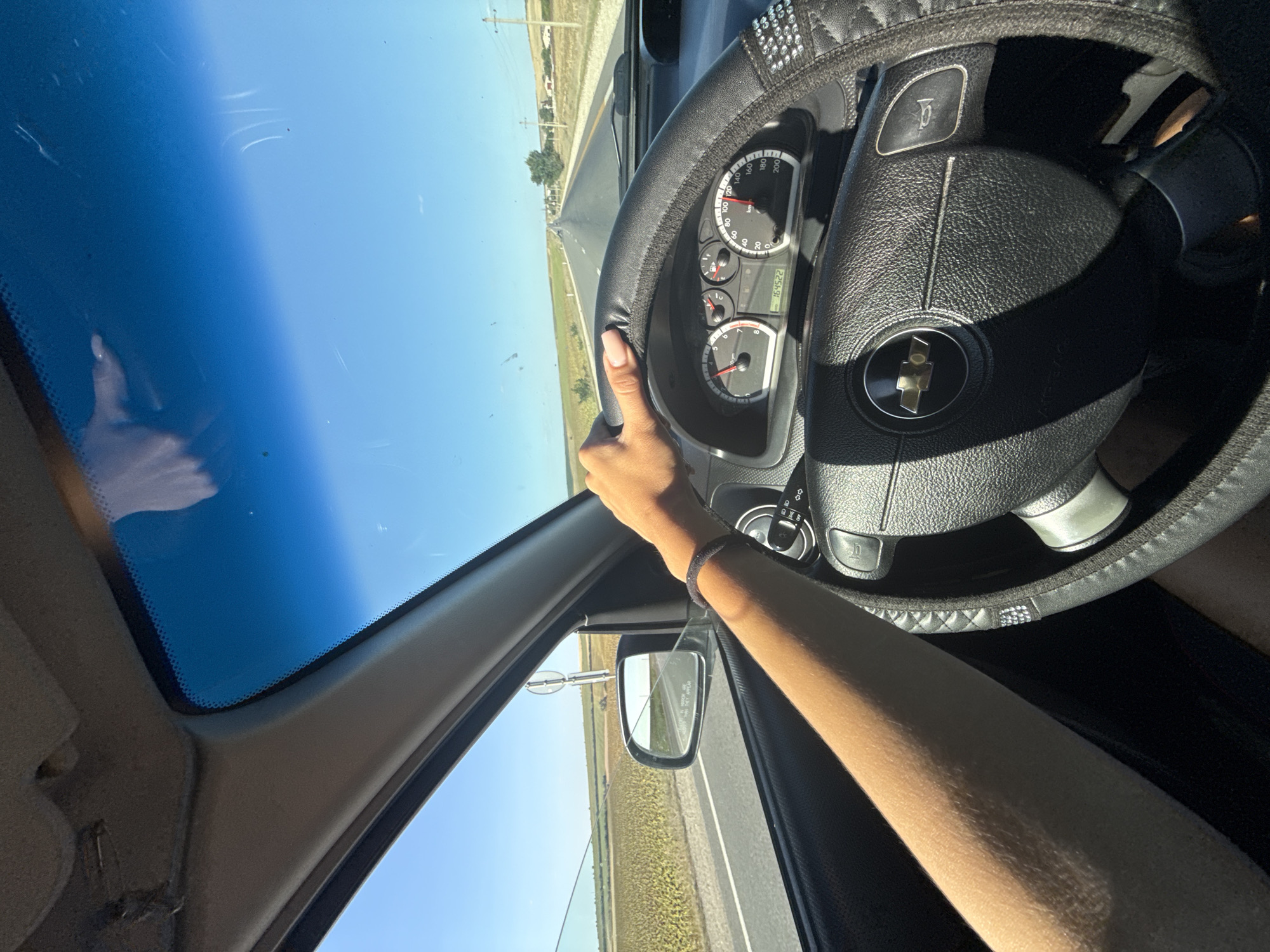 The image shows a first-person perspective of someone driving a car on a sunny day, with a clear blue sky and a road stretching into the distance. The car's interior is visible, including the steering wheel, dashboard with a speedometer, and a side mirror. The driver's arm and hand are on the steering wheel. Fields of crops, possibly sunflowers, are visible on either side of the road, suggesting a rural or semi-rural setting. The location is specified as the city of Comrat, Moldova.