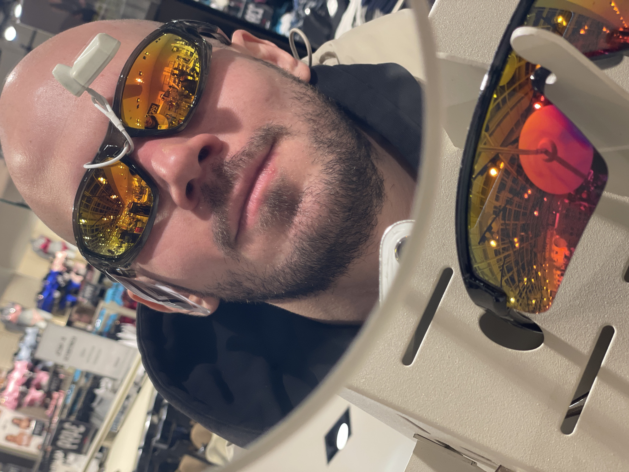 A man with a shaved head and a short beard is trying on sunglasses in a store. The sunglasses he is wearing have a white security tag attached to the left lens. The reflection in the lenses reveals a store interior with shelves and other merchandise. Additional sunglasses are displayed on a white perforated rack beside him. The location is likely a retail store in Krasnoyarsk, Russia, given the reference prompt.