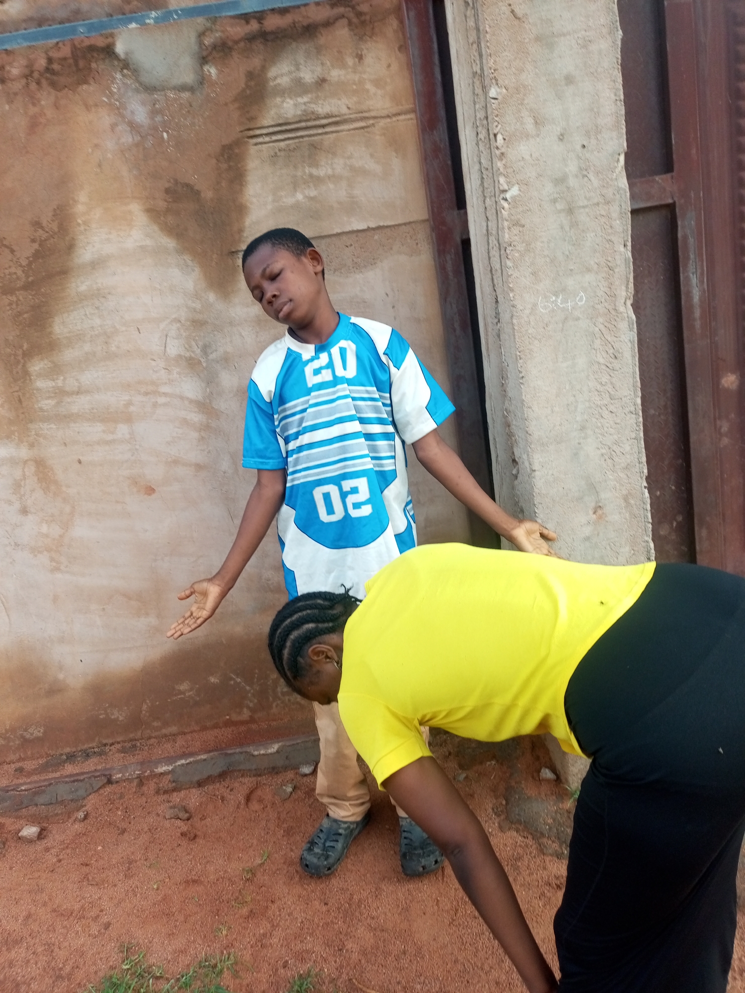 The image captures two young individuals in an outdoor setting, likely in Tempure, Nigeria. A young boy, wearing a blue and white jersey with the number "20" and "02" visible, stands with his arms outstretched and head tilted back, eyes closed, and a relaxed expression. To his right and slightly in front of him, a second individual, seen from the back, is bending over, wearing a bright yellow t-shirt and black trousers. The background consists of a textured, brownish-tan wall with some stains, and a concrete pillar with markings. The ground is covered in reddish-brown soil and a few patches of sparse green grass. The overall scene appears to be casual and perhaps depicts a moment of play or interaction between the two.