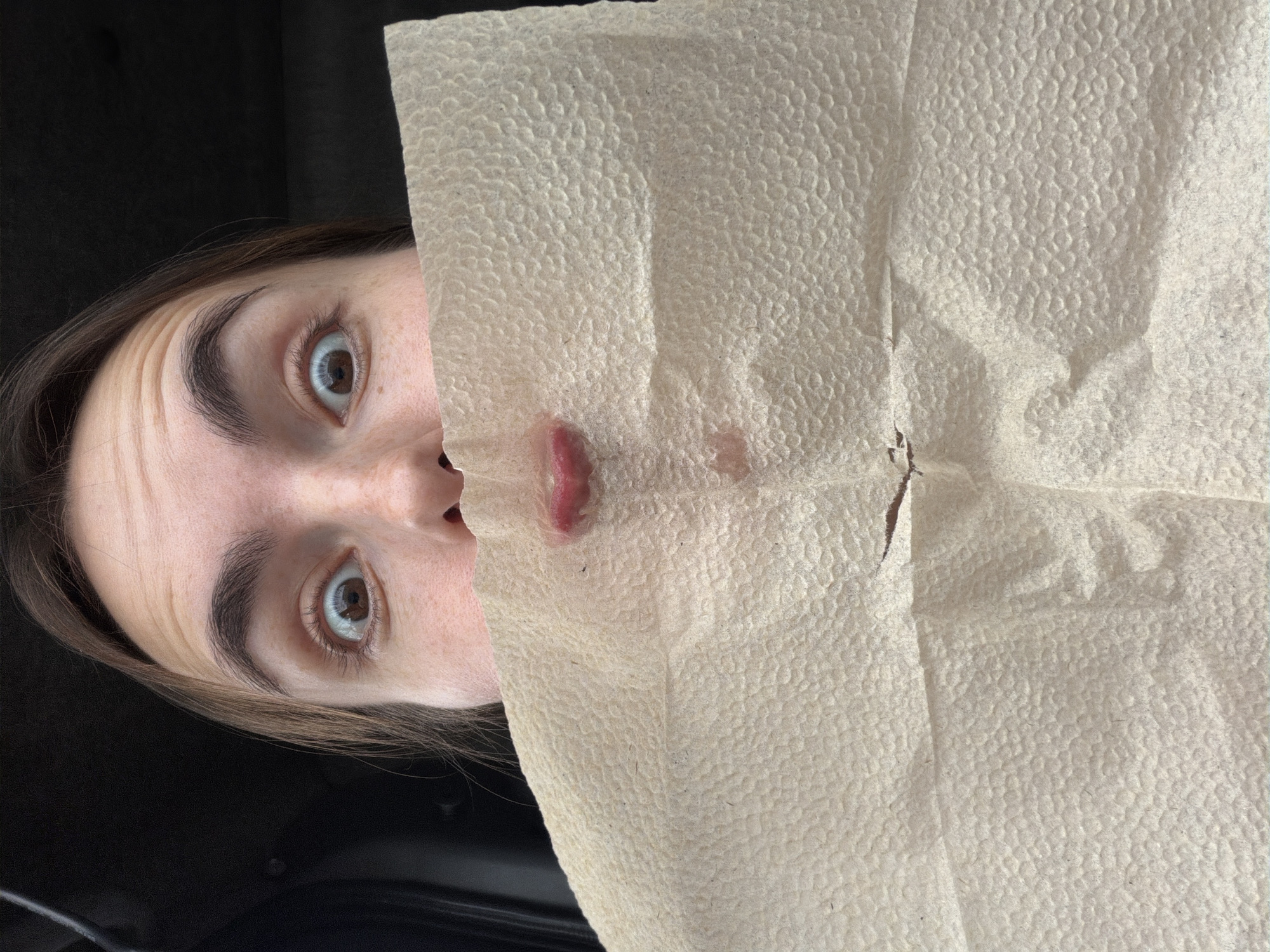 A close-up, eye-level shot captures a person, likely female, partially concealed by a textured, light-colored paper towel. The person's face is positioned on the left side of the frame, with the paper towel covering most of their nose and lower face. Their wide, expressive brown eyes and eyebrows are visible, suggesting an emotion of surprise, playfulness, or being startled. A small portion of their mouth, appearing slightly open, and a reddish stain or residue are visible on the paper towel near the lip. The background is dark and indistinct, resembling the interior of a car. The image is taken in Izhevsk, Russia. There is no visible text.