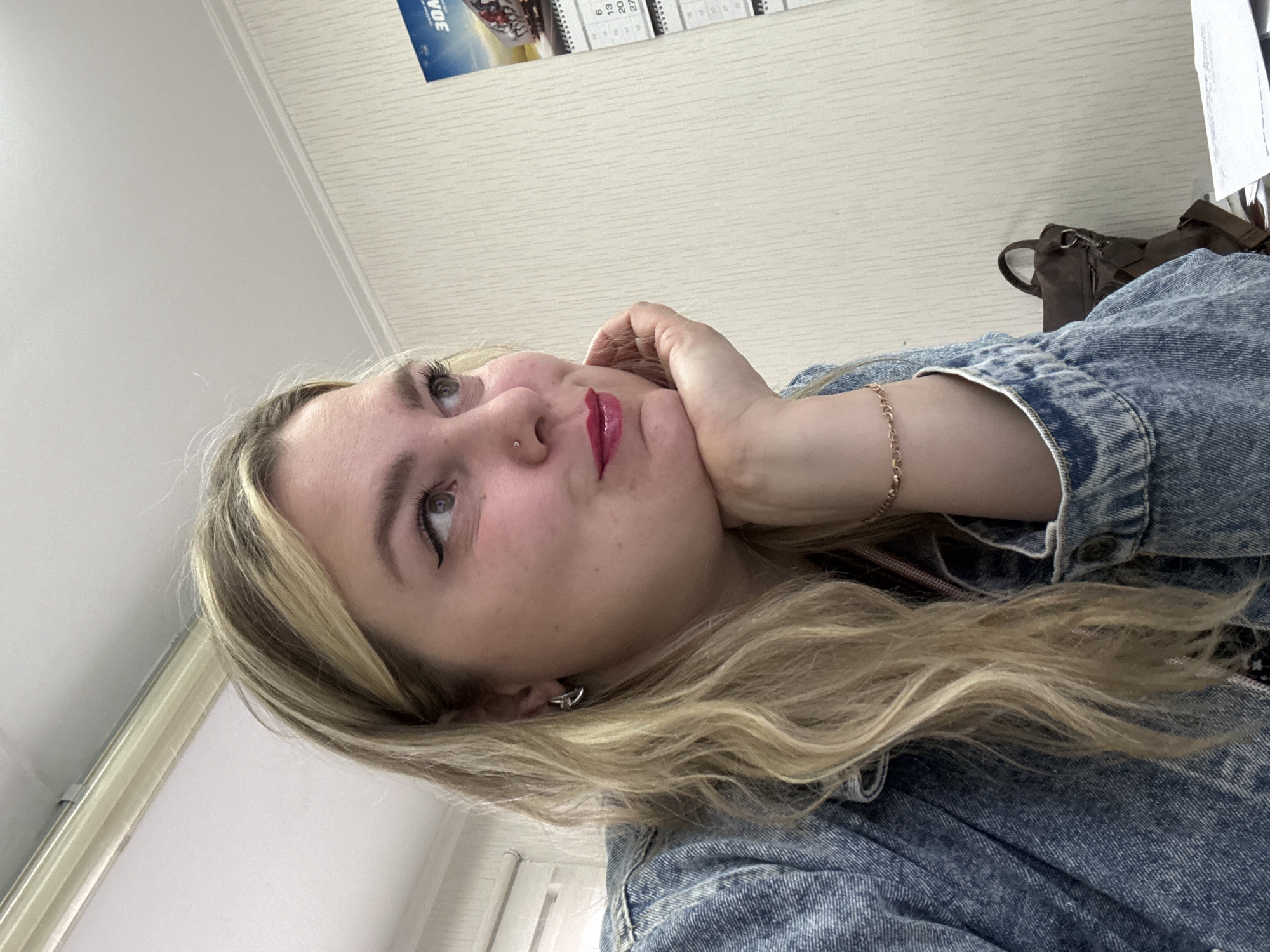 A woman with blonde hair is leaning her face on her hand. She is wearing a denim jacket. The image appears to be taken indoors with a calendar and bag visible in the background. The location is not discernible from the image.