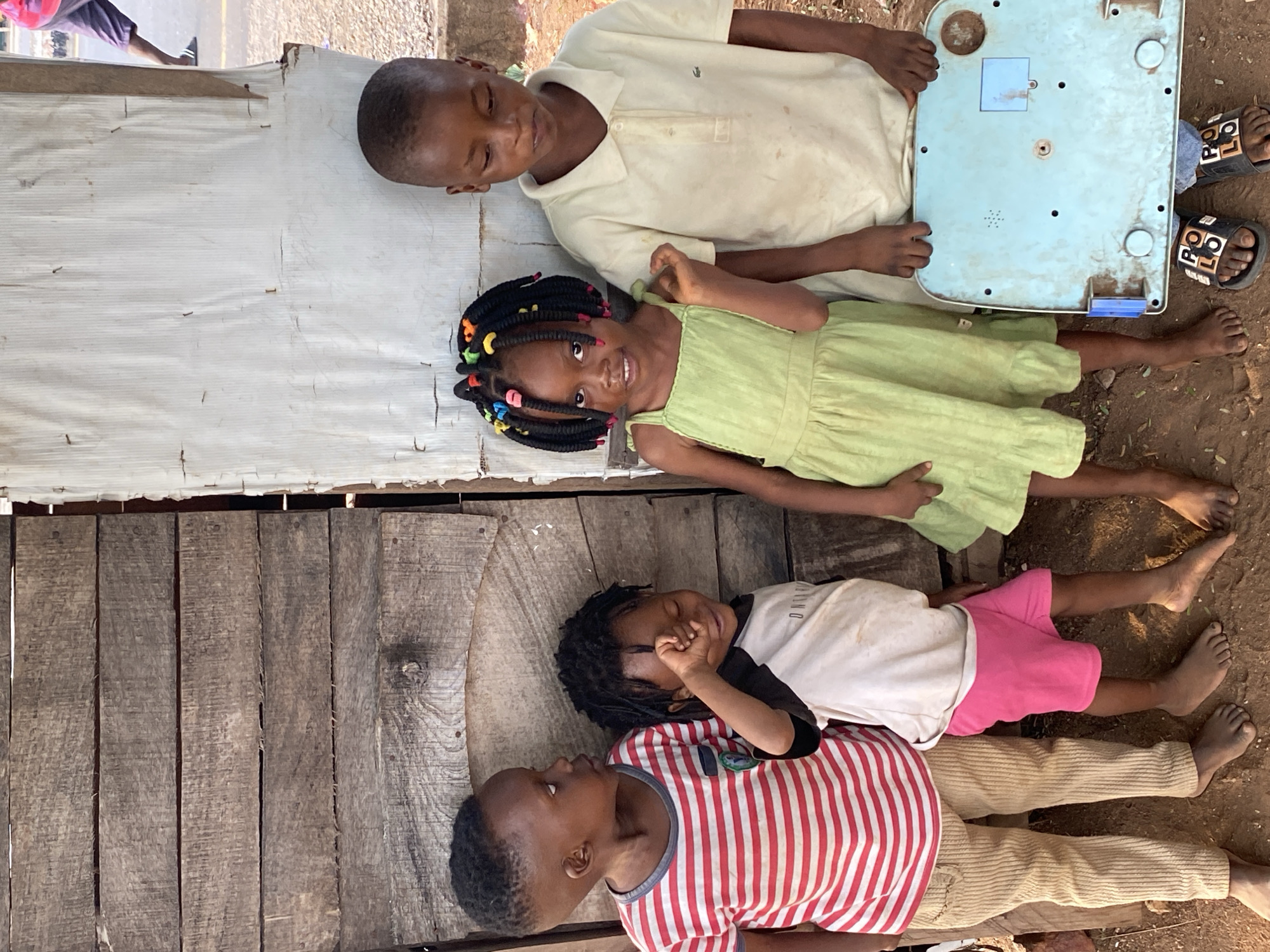 The image shows four children lying on the ground in Adankolo, Nigeria. The ground is dirt and wood planks. From top to bottom: a boy in a white polo shirt holds the underside of a light blue object; a girl in a green dress with colorful beads in her hair smiles; a child in a white shirt and pink shorts has their eyes closed; a child in a red and white striped shirt looks at the child in the white shirt. A pair of sandals rest near the boy holding the blue object.