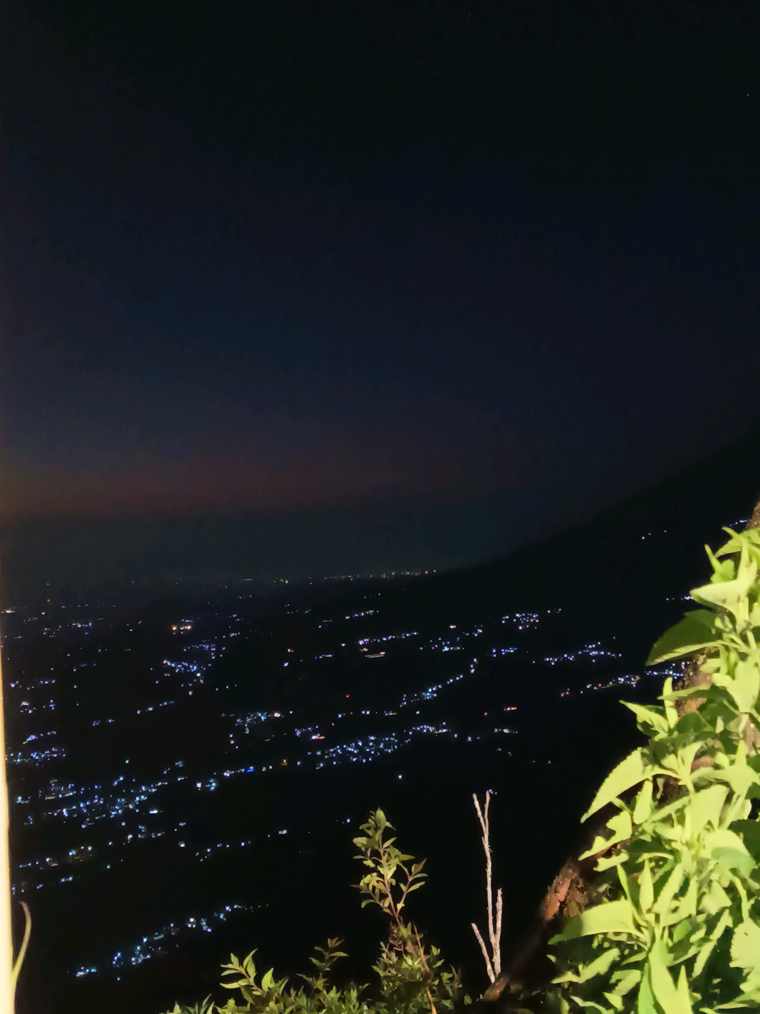 The image is a nighttime view of a city spread out in a valley from a high vantage point. The city lights are a prominent feature, appearing as scattered white and blue dots of varying intensity, suggesting a densely populated area. The lights form patterns that indicate streets and clusters of buildings. In the foreground, on the right side of the image, there are lush green plants with broad leaves, some of which are illuminated by an unseen light source. The background shows a dark, mountainous silhouette against a dimly lit sky, with a subtle gradient of color that might indicate the last vestiges of twilight. The overall impression is one of a serene, expansive cityscape at night, viewed from a natural overlook. There are no people or discernible human activity visible in the image. The location is identified as Keditan, Indonesia, suggesting this is a view overlooking that area.