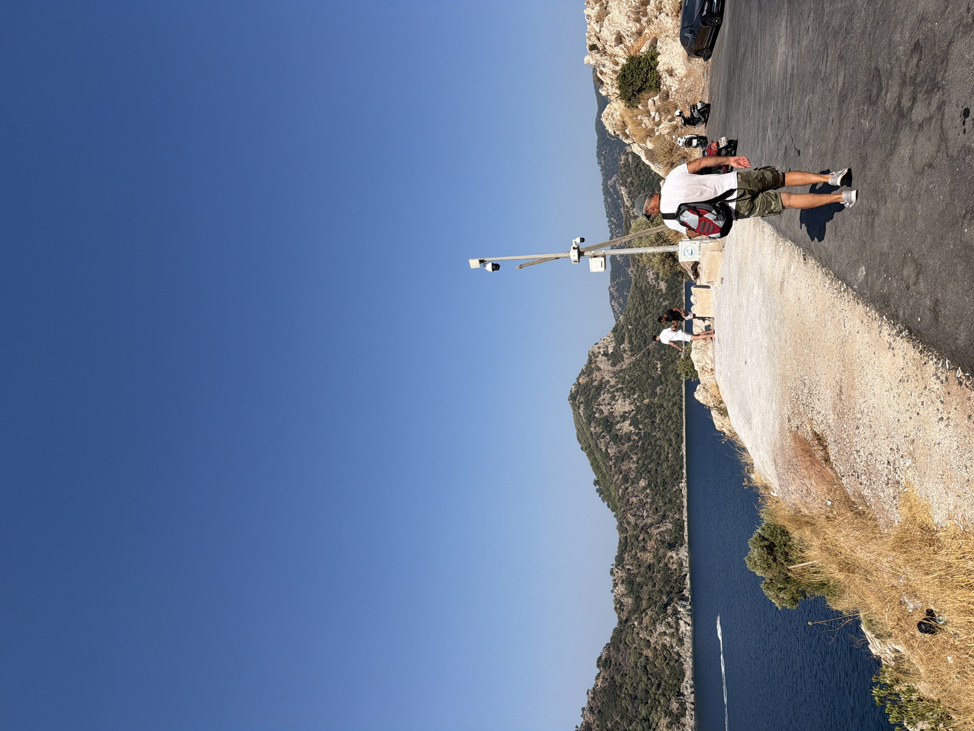 The image depicts a scenic view of Marmaris, Turkey, overlooking a bright blue sea and lush green mountains. In the foreground, a man with a backpack walks along a paved road. He is wearing a white t-shirt, camouflage shorts, and grey sneakers. Behind him, several motorcycles and a black car are parked. Further down the road, a group of people are standing on a concrete walkway, enjoying the view. The walkway is lined with dry grass and rocky terrain. In the distance, a boat is seen cruising on the water, leaving a white wake behind it. The sky is a clear, vibrant blue, characteristic of a sunny day in Marmaris. A security camera pole with multiple cameras is also visible near the walkway.