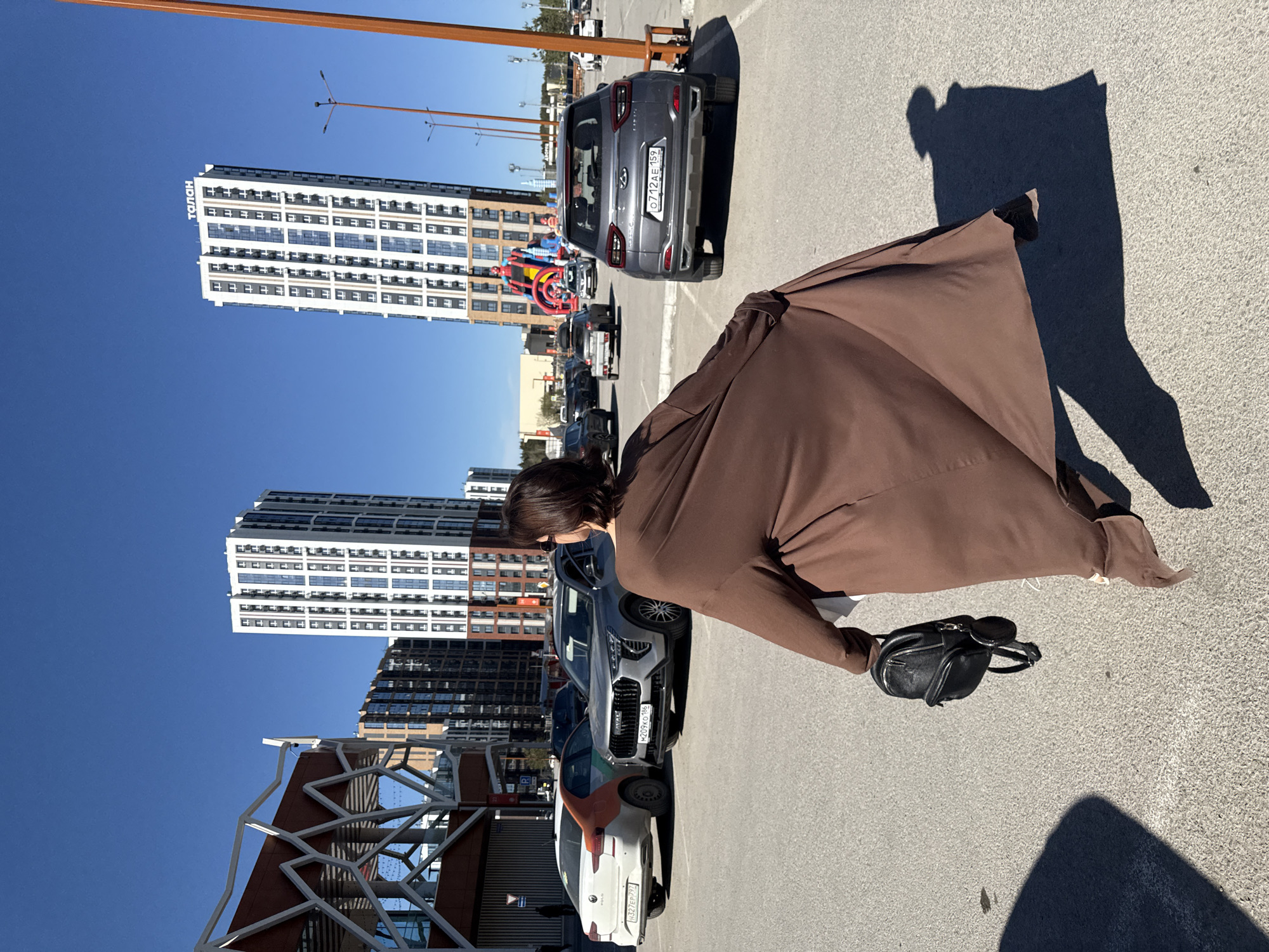 The image shows a person in a brown coat holding a small black purse. The scene appears to be set in a parking lot. In the background, there are two tall buildings, and several cars parked in the lot. The license plates on the cars are Russian license plates. An inflatable castle with images of Spiderman and other superheroes is also visible. The buildings appear to be located in the city of Perm, Russia.
