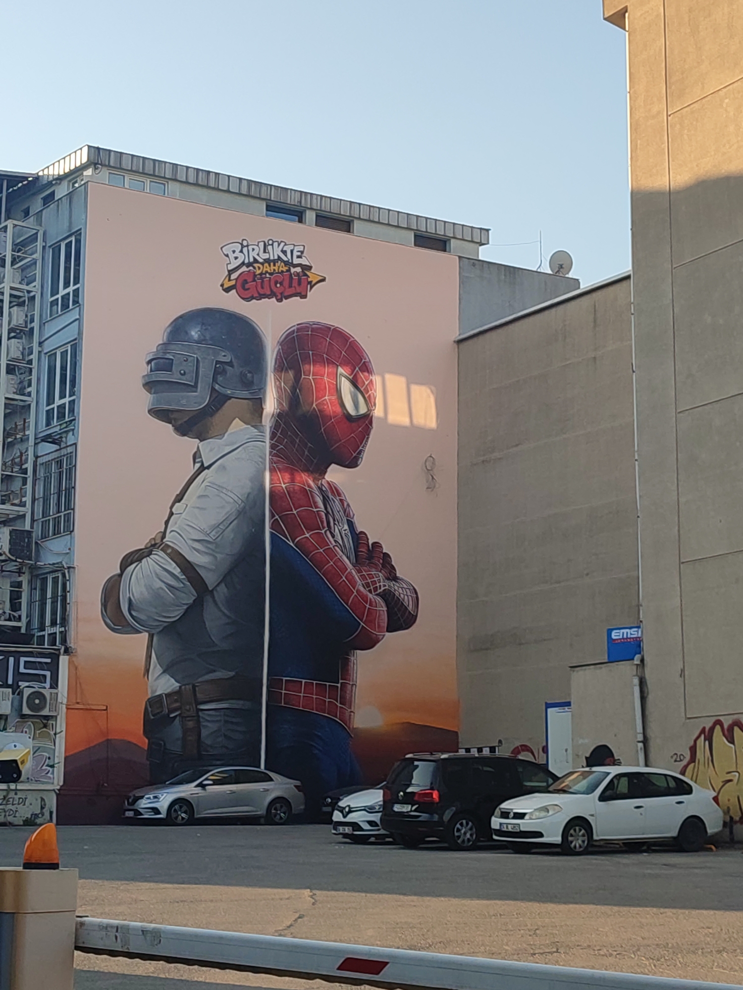 A mural is painted on the side of a building in Kadıköy, Turkey, depicting two characters: one in a helmet resembling a PUBG character, and the other in a Spider-Man costume. They are positioned back-to-back with their arms crossed. The mural is set against a backdrop of a sunset over a landscape. In front of the building, several cars are parked, including a silver sedan, a black SUV, and a white sedan. The area appears to be an outdoor parking lot or street. The sky is clear and blue. The building itself has multiple windows and air conditioning units visible. There is a street barrier in the foreground. Graffiti is present on some of the surrounding walls. A blue sign with white text reading "EMSI" is visible on a smaller building to the right.