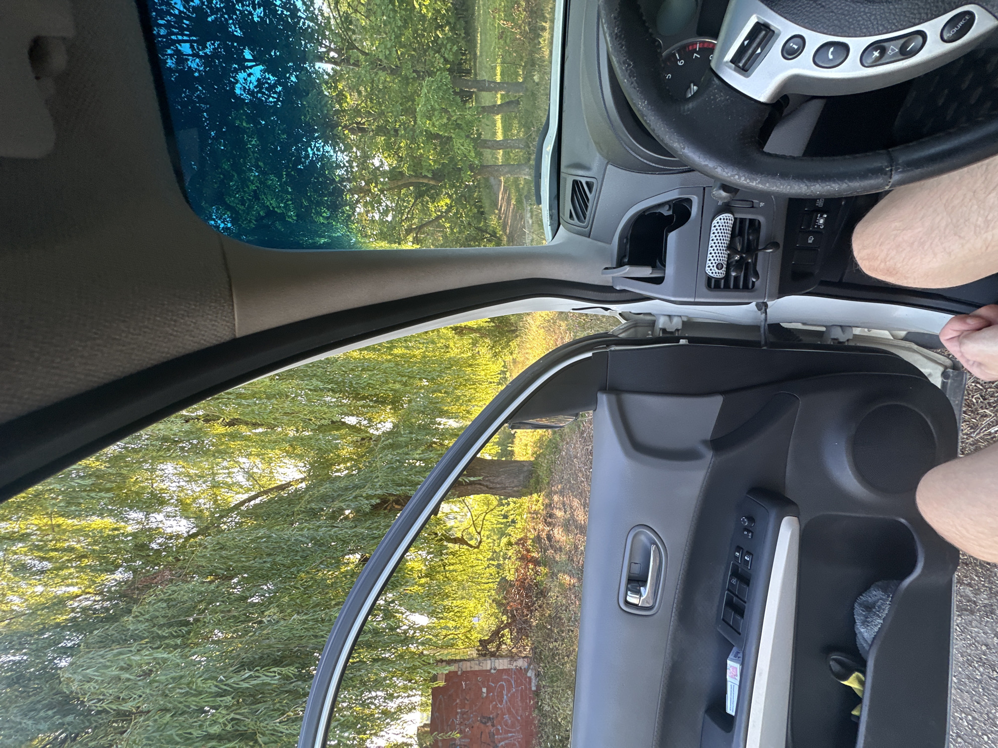 The image shows the interior of a car parked outdoors. The driver's side door is open, revealing a view of a rural or semi-rural environment with trees and vegetation bathed in sunlight. Inside the car, the dashboard, steering wheel, and door panel are visible. A person's legs are visible in the lower part of the frame, indicating their presence in the driver's seat. The scene appears to be one of rest or waiting, with no active movement depicted.