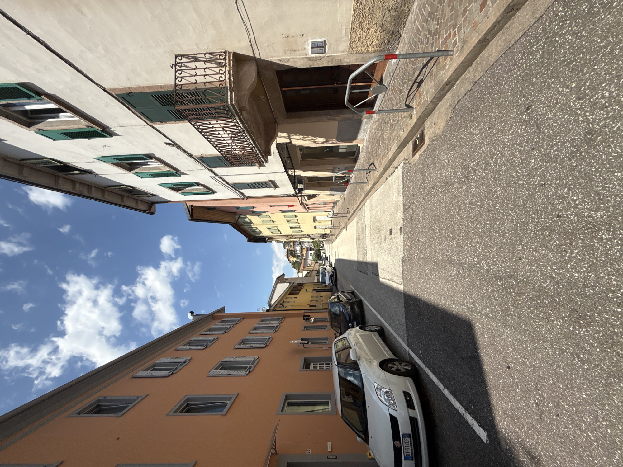 The image shows a narrow street in a European city, likely Italy, based on the architecture and the presence of a license plate on the white car that reads "DT-514DF" which is an Italian plate format. The street is lined with colorful buildings with balconies and shutters. The buildings on the left are painted orange, while those on the right are cream and yellow. The road is paved with asphalt and cobblestones, and there are parked cars on the left side of the street. The sky is blue with some white clouds. The scene appears to be taken during the daytime. The street is well-lit by the sun, casting shadows from the buildings and cars onto the road. There are no people clearly visible in the image. A white Suzuki Swift is parked on the left side of the street. A black car is parked behind it. Further down the street, a white van and other cars are visible. There are bollards along the sidewalk on the right side of the street. The buildings appear to be a mix of residential and commercial spaces, with shops visible on the ground floor of some buildings. The overall impression is of a quiet, picturesque street in a historic town.