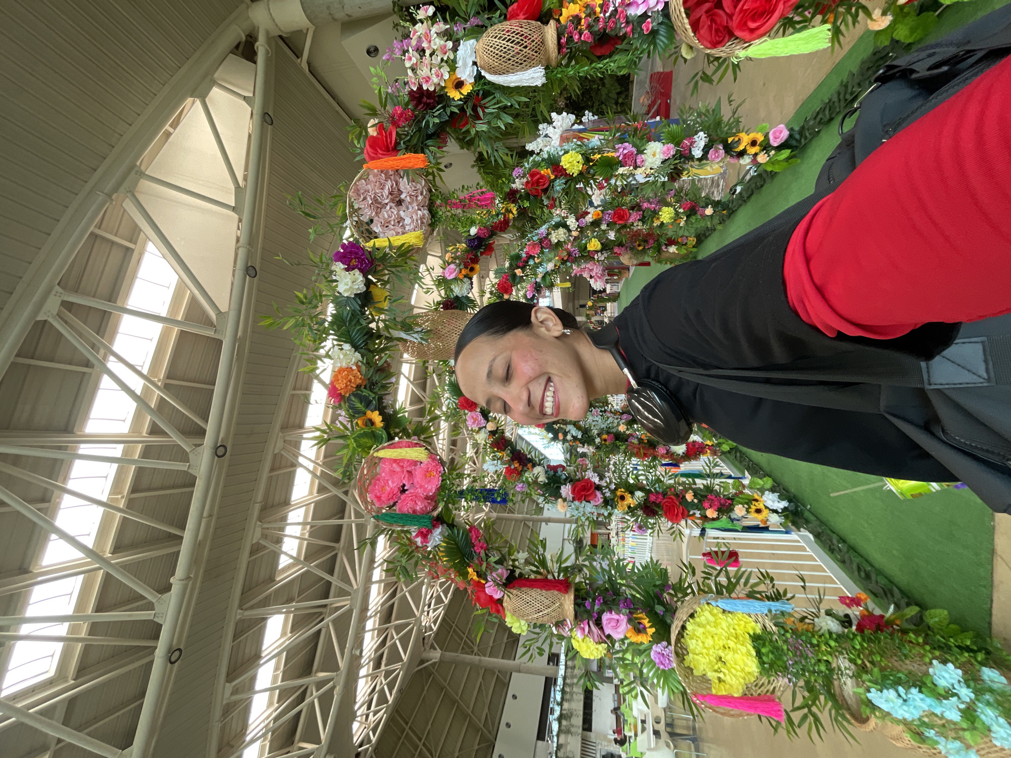 A woman takes a selfie in front of a floral archway adorned with colorful artificial flowers and decorative baskets, likely at a festive event in Pampatar, Venezuela. The archway is set up outdoors, possibly in a public space or venue, with a high, industrial-style ceiling visible in the background. The woman is smiling broadly, her eyes closed, and she is wearing a black shirt with a red sleeve and a black bag slung over her shoulder. The ground in front of the archway is covered in green artificial turf. In the background, tables and chairs of a restaurant or seating area can be seen, as well as some white fencing. The overall impression is one of celebration and joy.