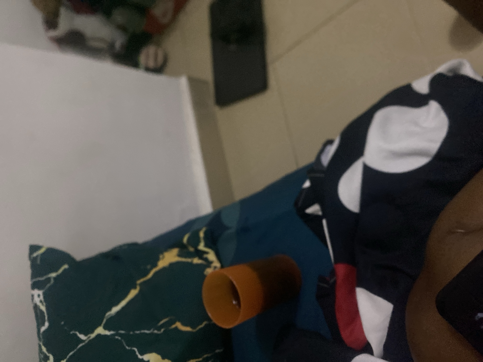 The image shows a dimly lit bedroom in Howayi, Nigeria. A dark green pillow with a gold marble pattern is visible on the left side of the frame, placed on a teal-colored surface, likely a bed or sofa. A translucent orange cup, partially filled with a dark liquid, rests on the teal surface next to the pillow. In the foreground, a patterned fabric, primarily black with white circles and red and white stripes, is draped across the lower part of the image, suggesting clothing or bedding. The background features a light-colored wall and a beige tiled floor, with a dark rectangular object, possibly a mat or a book, placed on the floor to the right. The lighting is low, creating shadows and a sense of an interior space, likely a bedroom. There are no visible people in the frame.