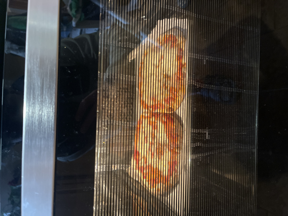 The image captures a close-up, top-down view of food, likely a pizza or flatbread, being cooked inside an appliance such as an oven or microwave. The food item, characterized by its reddish-orange sauce and melted, yellowish cheese, is partially obscured and distorted by a series of vertical, dark slats, which appear to be part of the appliance's door or protective screen. The food itself shows signs of being well-cooked, with some browning visible on the cheese and crust.

To the left of the slatted view, a prominent, brushed metallic vertical bar is visible, suggesting part of the appliance's handle or frame. The surrounding areas are dark and reflective, showing vague reflections of the kitchen environment and possibly a person's hand or arm. The overall setting is an indoor kitchen. There is no visible text in the image. The unique perspective through the slats creates a striped effect over the food.