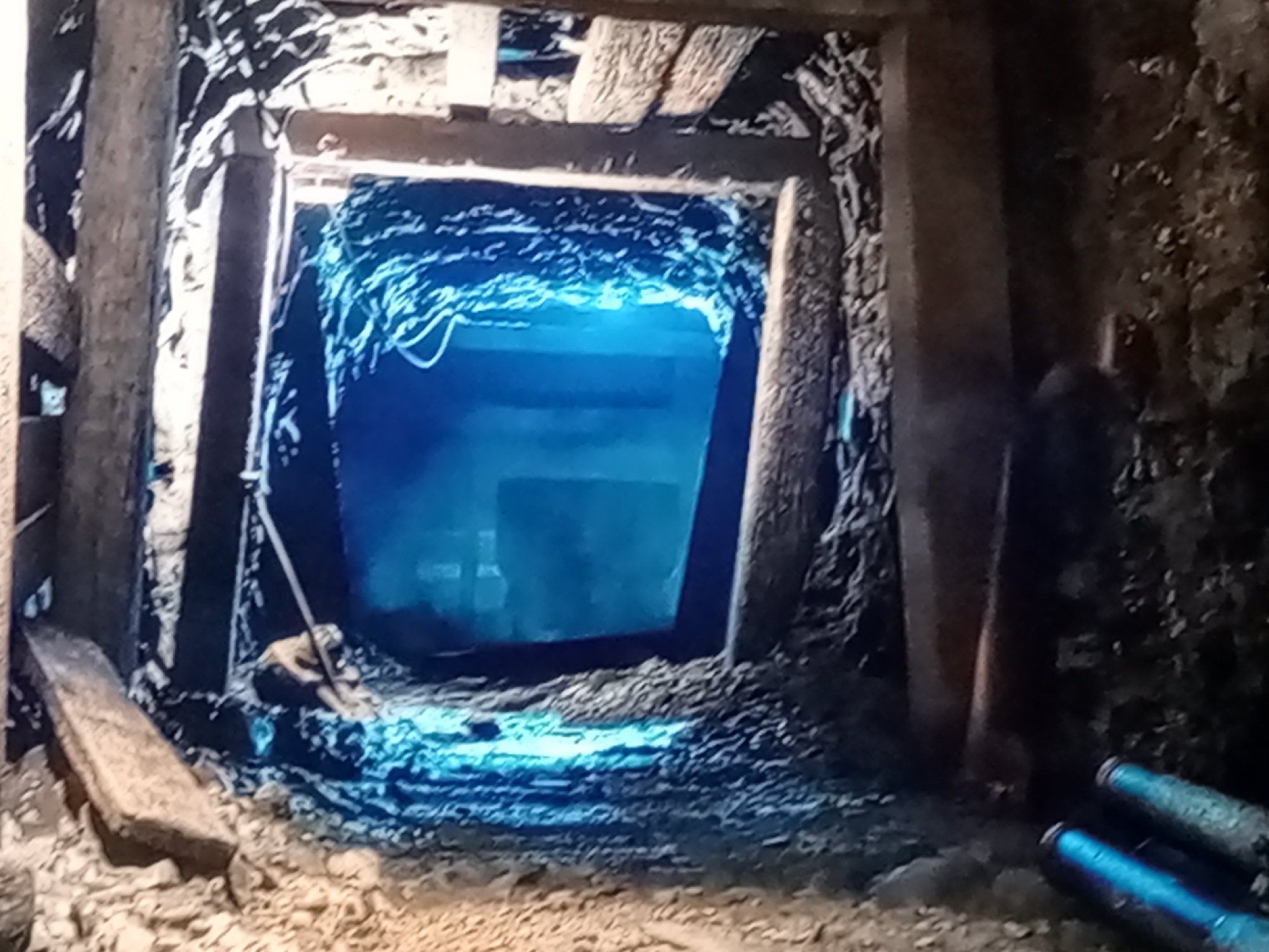 The image depicts a dimly lit, rough-hewn tunnel or mine shaft. Wooden beams support the ceiling and walls, which appear to be made of rock and dirt. The tunnel opens into a larger, brighter space illuminated by a vibrant blue light, creating an otherworldly atmosphere. The ground is covered in loose rubble and debris. In the foreground, a person is partially visible, hunched over or resting near a pile of rocks. Two cylindrical objects, possibly pipes or tools, are visible in the lower right corner. The overall impression is one of a subterranean environment, potentially a working mine or an abandoned one.
