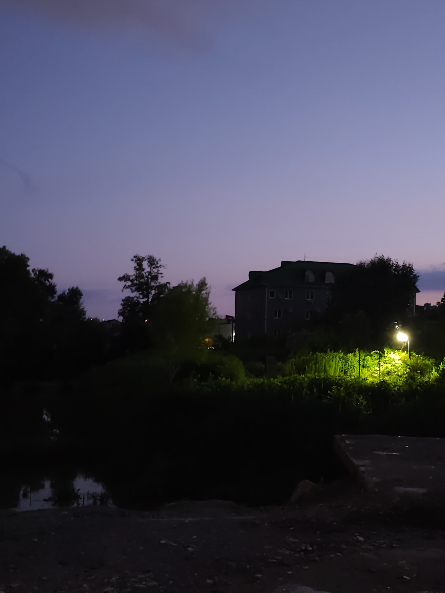 The image depicts a dusky twilight scene with a purple-hued sky above. In the mid-ground, a multi-story building with a green roof is silhouetted against the sky, surrounded by dark foliage. To the right of the building, a bright street lamp illuminates lush green vegetation. In the foreground, a dark body of water, possibly a pond or river, reflects the dim sky and surrounding trees. The ground in the immediate foreground appears to be rough, possibly gravel or dirt, with some scattered stones. The overall impression is one of a serene, somewhat rural or suburban evening.