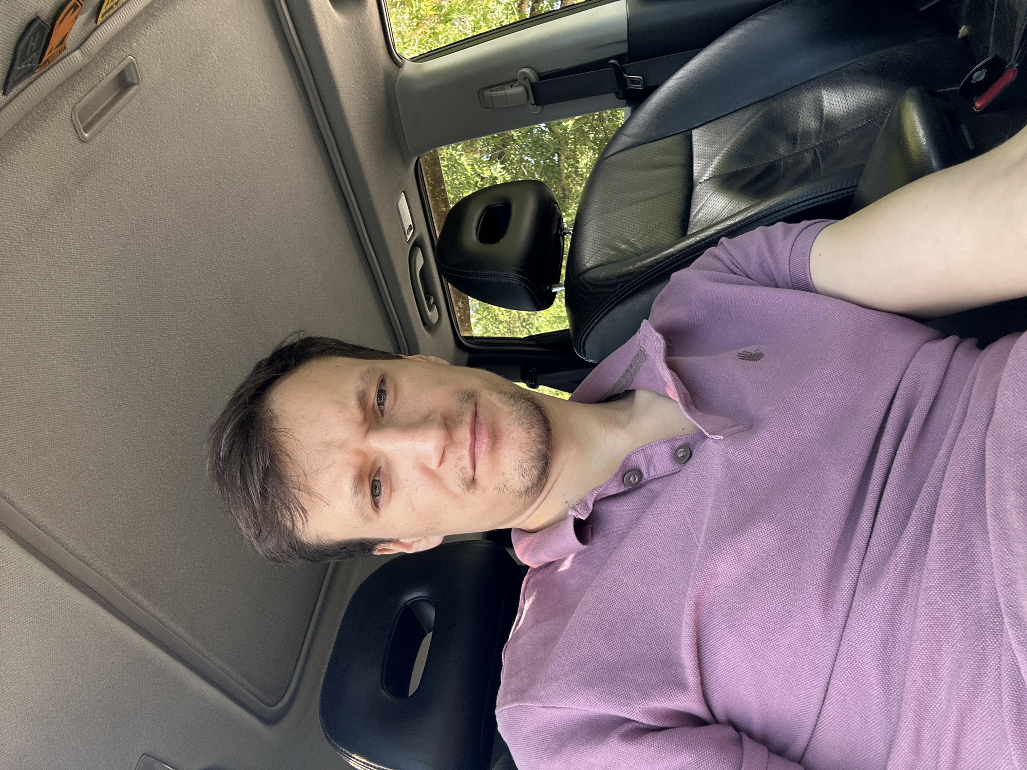 A man is sitting in the driver's seat of a car, facing the camera. He is wearing a light purple polo shirt. The car's interior is visible, including the black leather seats, the sunroof, and the dashboard. Outside the car, trees and foliage are visible through the windows. The lighting suggests it is daytime. The location is the city of Makiv, Ukraine.