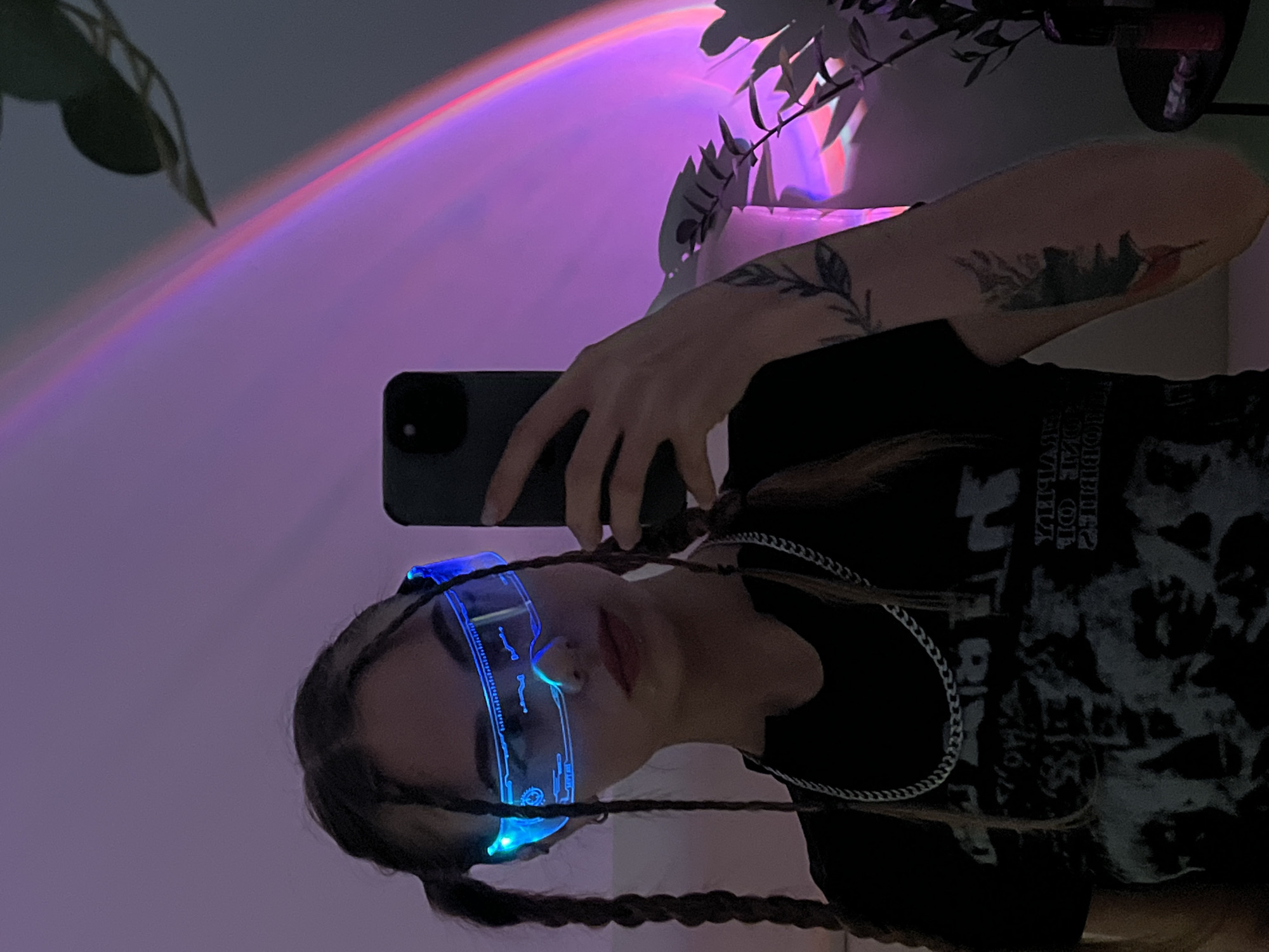 A young person with long, braided hair is taking a selfie with a dark-colored smartphone. The individual is wearing black clothing and cyberpunk-style glasses with blue lights. They also have tattoos on their arm. The background is a soft pink and purple hue, possibly from a light fixture or filter. There is a plant in the background. The location appears to be indoors. The city of Фоки, Russia cannot be confirmed with the current information.