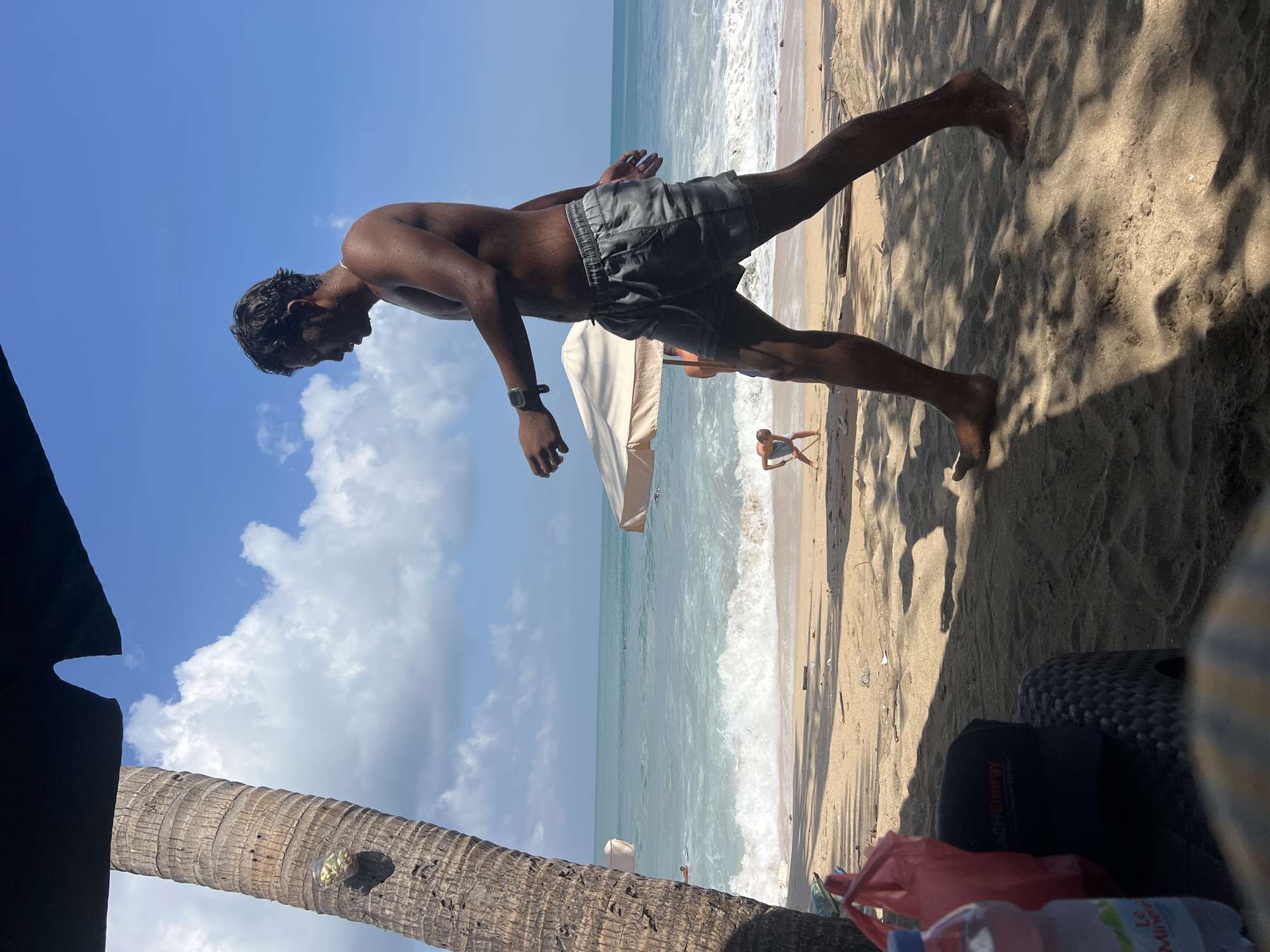 In Kuta, Indonesia, a man with dark hair, wearing grey patterned shorts and no shirt, walks to the right along a sandy beach. He wears a watch on his left wrist. The ocean with white waves is behind him, under a bright blue sky with scattered clouds. A palm tree stands to the left of the frame. In the distance, another person, possibly a child, is visible near the water, holding a stick. Two large beach umbrellas are set up on the sand. In the foreground, to the left, a plastic water bottle and a red plastic bag are on a dark woven surface.