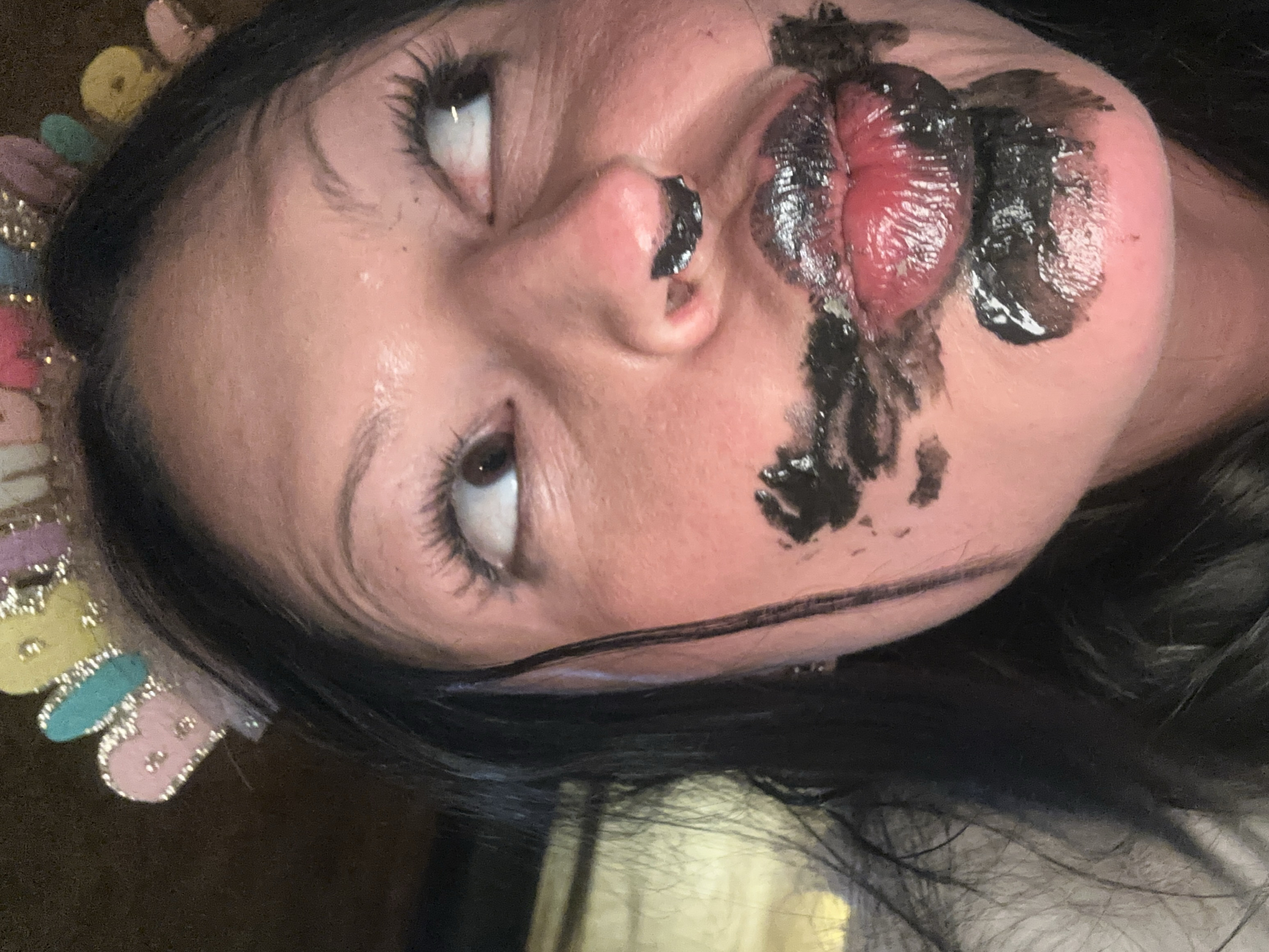 A close-up portrait shows a person's face angled upwards and to the left. The individual has dark hair framing their face and dark eyes, with prominent eyelashes, looking towards the top left of the frame. A thick, dark, glossy substance, resembling black paint or heavy makeup, is smeared extensively around their mouth, covering parts of the upper lip, lower lip, chin, and cheek, as well as a small spot on the tip of the nose. The lips beneath the substance appear very red, shiny, and somewhat swollen or accentuated. A distinct, thin, horizontal black line is drawn across the lower cheek and chin area, separate from the smudged substance. On the left side of the head, partially visible against a dark background, are colorful, glittery, and possibly fabric-like decorative elements in shades of yellow, teal, and pink, suggesting a headband or crown. The person's skin shows some slight sheen, possibly from moisture or product. The expression conveyed by the upturned eyes could be one of playfulness, surprise, or theatricality. No text is visible in the image.