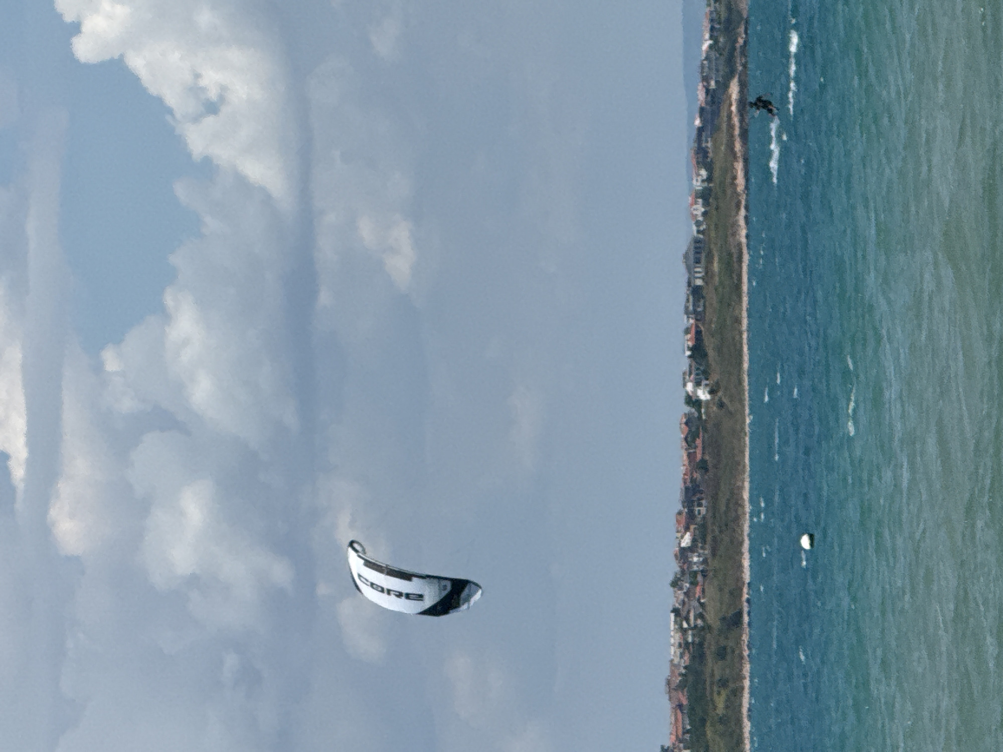 A bright, cloudy sky is visible above a body of turquoise water. A white and black kite, with the brand name "CORE" visible in black lettering, is suspended in the air. In the distance, a densely populated coastline is lined with red-tiled roofs and green foliage. A lone buoy floats in the water. A person on a surfboard is visible in the water on the right side of the frame, performing a kitesurfing maneuver, creating a spray of water behind them. This scene likely depicts a day of kitesurfing in or near Sozopol, Bulgaria, given the characteristic coastline and clear waters.