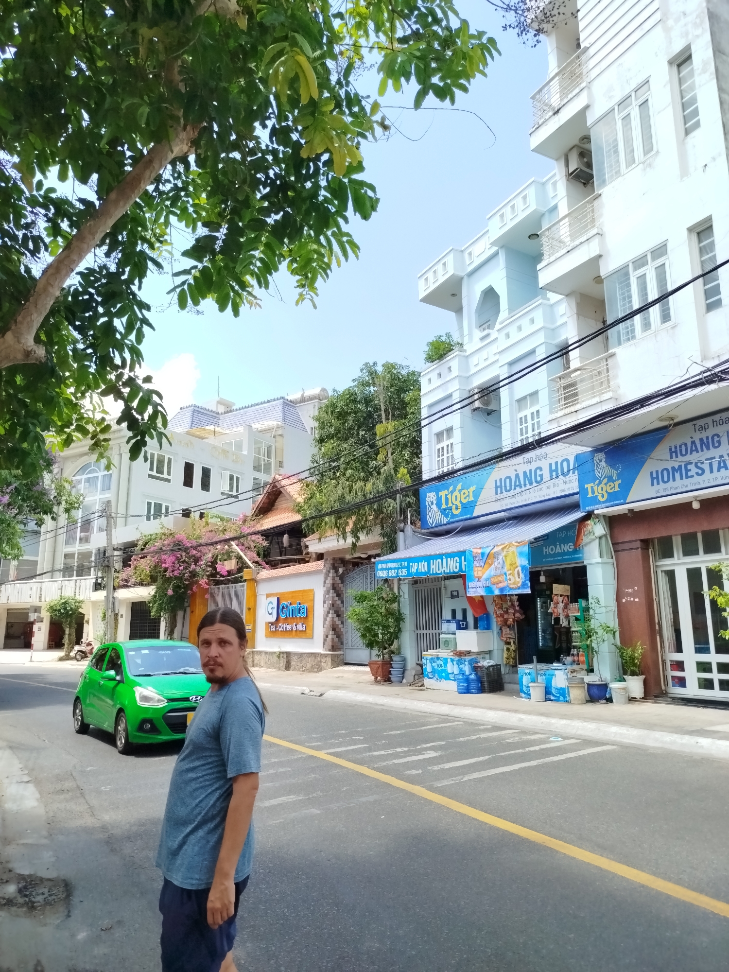 The image captures a street scene in what appears to be a residential area of Ho Chi Minh City, Vietnam. A man stands in the foreground facing the camera, slightly off-center. Behind him, a bright green car is parked along the side of the road. A yellow line marks the lane divider on the asphalt road.
On the right side of the frame, a multi-story building is visible with a light blue façade and white trim. A storefront operates under the building, indicated by the "Tiger" beer advertisement and awning with signage reading "TẠP HÓA HOÀNG H." Various goods and bottled drinks are displayed outside the store.
On the left side of the image, there are other buildings, one with bougainvillea growing over the wall, and signage that reads "Ginta Tea - Coffee & Villa". A tree with dense foliage occupies the upper left corner of the frame. The sky is pale blue. The scene is bathed in daylight, suggesting a sunny day.