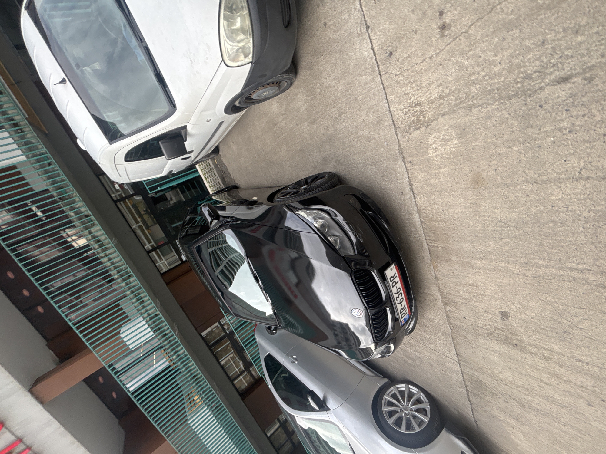 The image shows a street scene with three parked cars. A white van is parked at the top of the frame, followed by a black BMW, and a silver sedan parked below the BMW. The license plate of the BMW is visible and reads "RR-636-PR." The cars are parked on a gray concrete surface. To the left of the cars is a green metal railing with a building behind it. The location is likely in Tbilisi, Georgia.