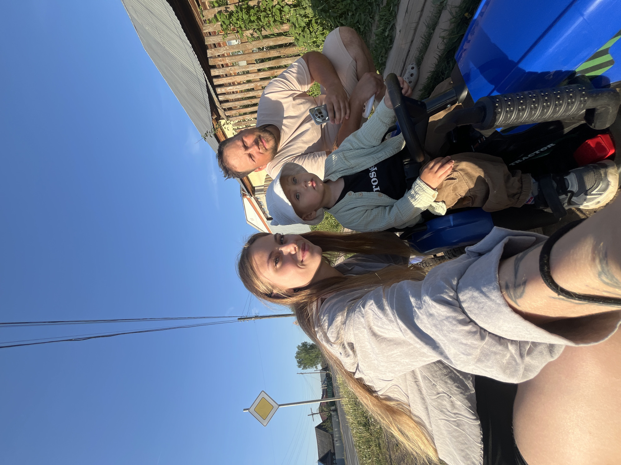 The image depicts a family of three taking a selfie in what appears to be a rural or suburban setting. A woman with long blonde hair and a grey shirt is holding the camera. Seated behind her is a man in a tan shirt, and a toddler wearing a light green sweater, dark shirt, and light-colored hat is sitting in front of the man on a blue plastic tractor toy. The background shows a clear blue sky, utility poles with wires, and a road sign. A wooden fence and some buildings can also be seen. Based on the location Фоки, Russia, the scene is likely in a residential area with a mix of houses and green spaces.
