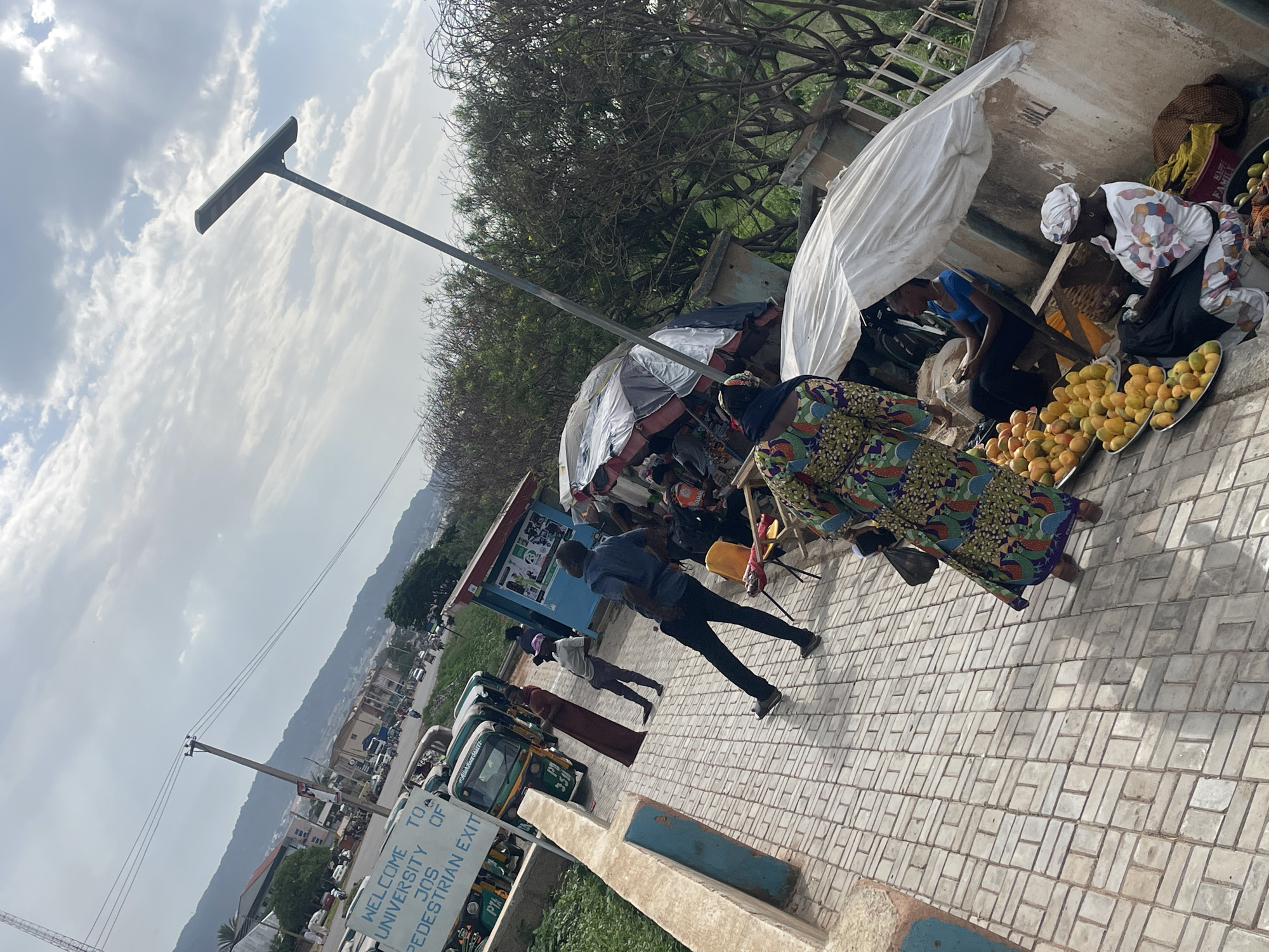 The image captures a vibrant outdoor scene near the University of Jos in Nigeria, oriented at an angle. The foreground features a paved walkway constructed from interlocking concrete blocks.
On the right, several street vendors are set up under makeshift shelters of white and gray tarpaulins. Prominently displayed on round metal trays on the ground are large quantities of bright yellow and orange fruit, likely mangoes. Two women appear to be working at these stalls; one in a white headscarf and patterned top is crouched over her merchandise, while another in a colorful patterned dress walks nearby. A person in a blue shirt is also seen attending to a stall behind the first vendor. A man in a dark blue shirt and black trousers is walking across the paved area in the center.
In the middle ground, a road is visible with several auto-rickshaws, commonly known as "Keke Napep" in Nigeria, parked or in transit. Text such as "Allahnasham" and "PD 933 JJI" is discernible on one of these vehicles. A tall, modern streetlight with a solar panel top extends upwards from the left side of the scene.
A white sign, partially visible on the left side of the road, clearly states "WELCOME TO A UNIVERSITY OF JOS PEDESTRIAN EXIT," providing a precise location cue. The background features scattered buildings and distant hills or mountains under a partly cloudy sky, characteristic of the Jos plateau. The bright, diffuse light suggests it is daytime, possibly mid-morning or afternoon, casting soft shadows. The scene depicts a common street market environment near a university entrance, bustling with daily commerce and pedestrian activity.