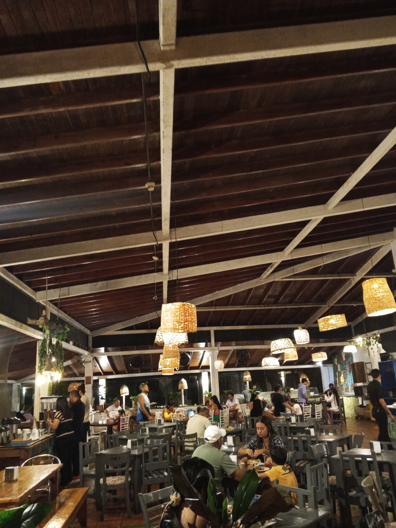 The image captures the interior of a restaurant. Many diners are seated at tables, interacting with each other or being served by staff. The restaurant has a high, wooden ceiling structure with white supports. Wicker lamps hang from the ceiling, providing illumination. The chairs and tables are mostly gray. The restaurant appears to be in Porlamar, Venezuela.