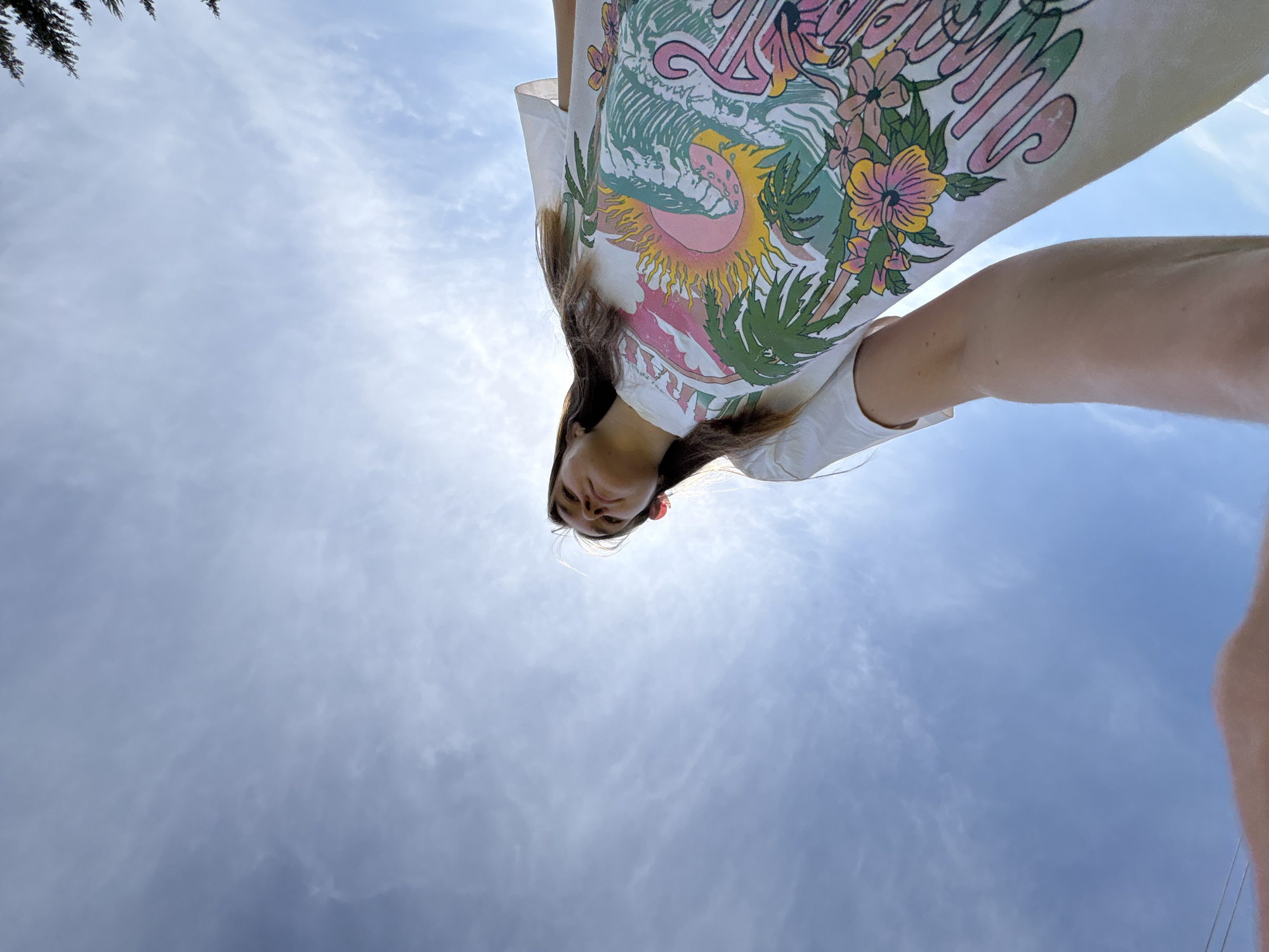 A low-angle shot of a fair-skinned person with long brown hair, wearing a white t-shirt with a colorful floral and sun-themed graphic. The person's face is visible, with a slight smile. The background is a cloudy sky with the sun shining through. The location is not discernible from the provided image and cannot be related to Sibir', Russia.