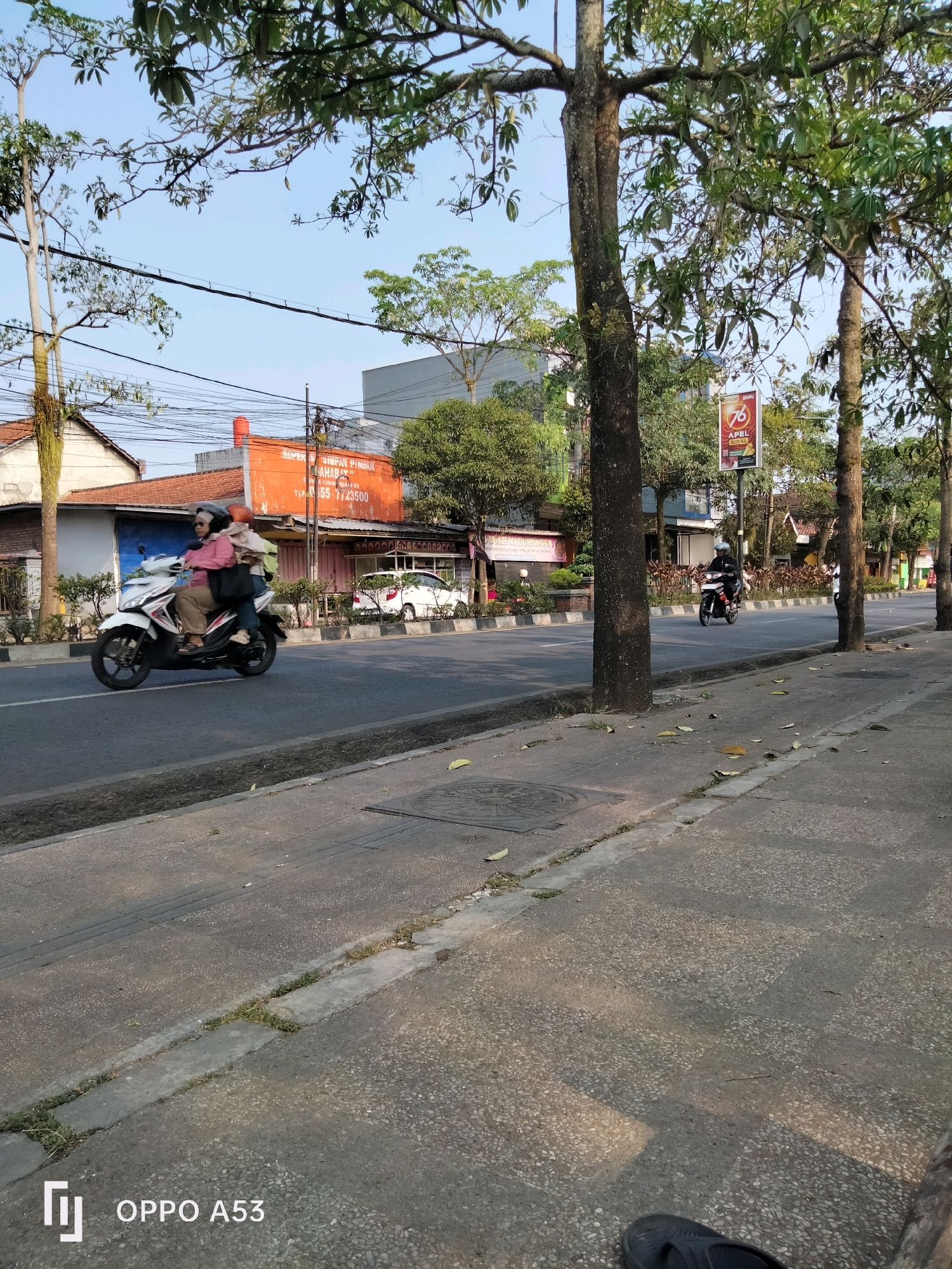 The image captures a street scene in Kelutan, Indonesia, with a focus on vehicular traffic and the surrounding urban environment. In the foreground, a paved sidewalk runs alongside a street. A man on a motorcycle wearing a helmet is visible in the middle ground, traveling in the same direction as the viewer. Further down the street, a white scooter is carrying two passengers, also with helmets, moving in the same direction. The street is lined with trees and buildings, some of which are commercial establishments. A bright orange building with signage is prominent on the left side of the street. The sky is clear and blue, suggesting daytime. The overall impression is of a typical day in a bustling Indonesian city.