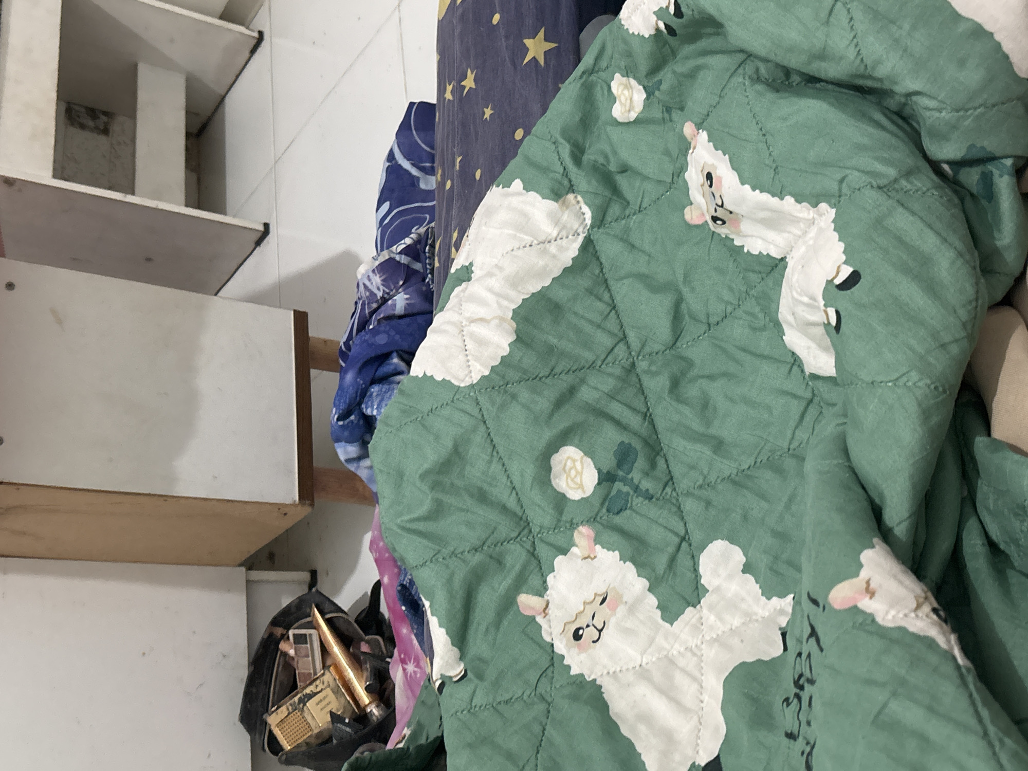 A close-up shot captures a green quilt with a pattern of white llamas and roses. The quilt is draped over a bed with a navy blue comforter featuring yellow stars visible underneath. To the left, a black makeup bag lies open, spilling out various makeup items including eyeshadow palettes and tubes of foundation. Behind the makeup bag, a portion of a wooden cabinet is visible. The floor appears to be tiled. The image is likely taken indoors, possibly in a bedroom in North Jakarta, Indonesia, given the context provided.