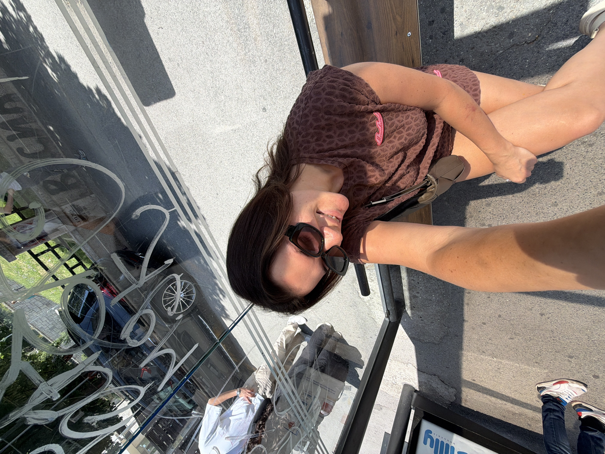 A selfie taken from a high angle shows a woman sitting on a bench at a bus stop. The woman is wearing sunglasses and a brown patterned t-shirt. She is smiling and looking at the camera. A man is visible in the reflection of the bus stop glass, wearing a white t-shirt and beige pants. The bus stop is located in Belgrade, Serbia, and the background shows a street with cars and trees. The lighting suggests it is a sunny day.