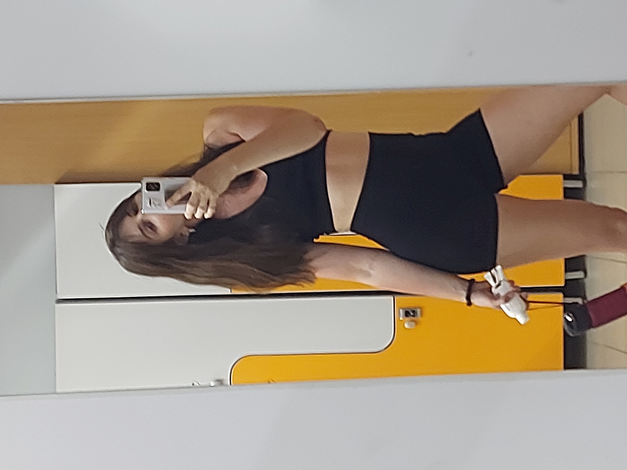 A woman is taking a selfie in a locker room. She is wearing a black sports bra and black shorts. She is holding a phone in her right hand. Her left hand is holding a white cloth or towel. The lockers are white and yellow. A purse is visible on the right. The location is presumed to be Chaikovsky, Russia, but no specific visual elements confirm this location.
