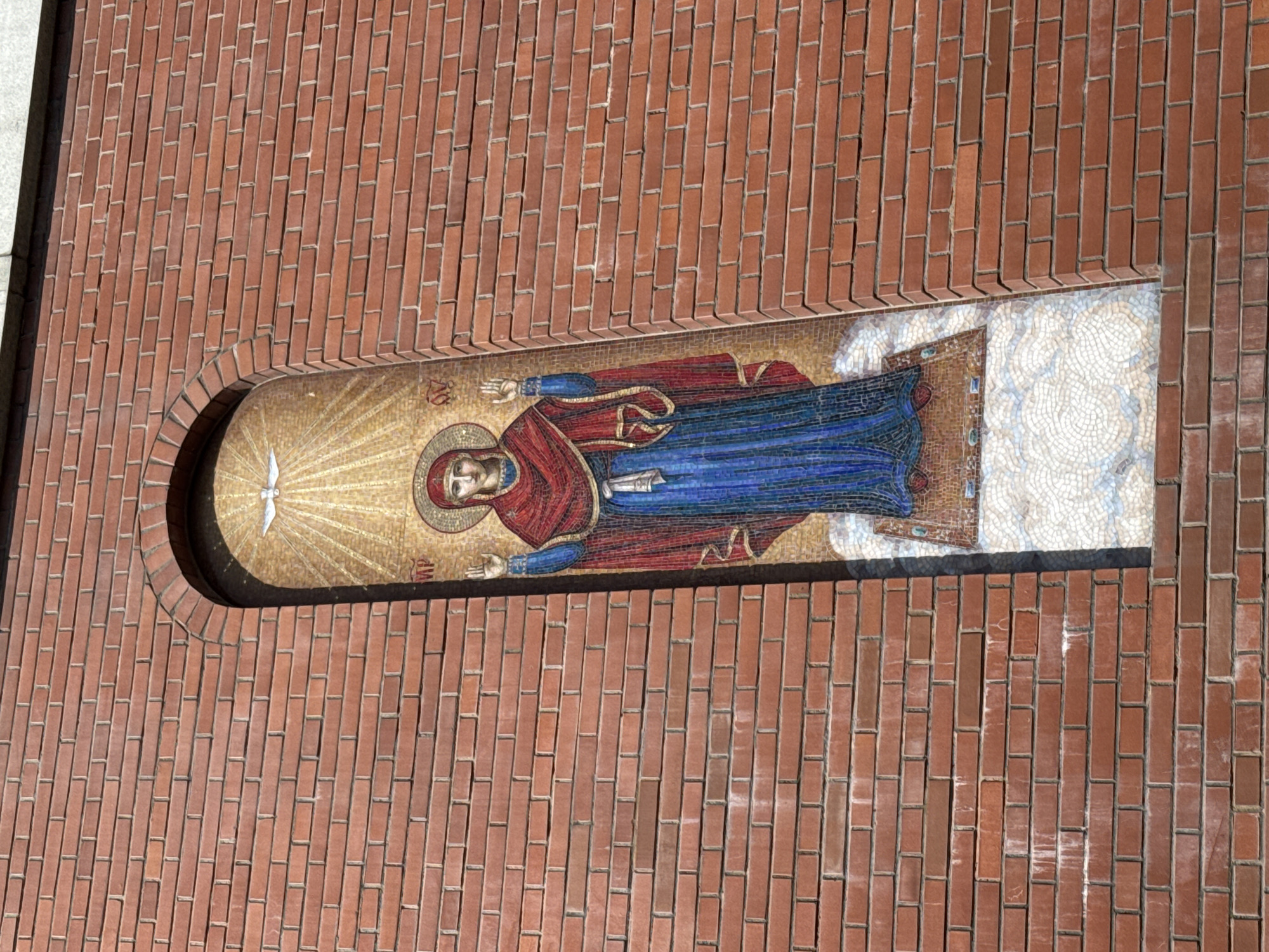 The image shows a mosaic of the Virgin Mary on the facade of a brick building, likely a church, in Sinyavino, Russia. The mosaic is set within a recessed arched niche. The Virgin Mary is depicted with outstretched arms, wearing a blue robe and a red veil. Rays of light emanate from above, and a white dove, symbolizing the Holy Spirit, is visible at the top of the niche. The surrounding brickwork is a common building material, suggesting an urban or suburban setting. The sunlight indicates it is daytime.