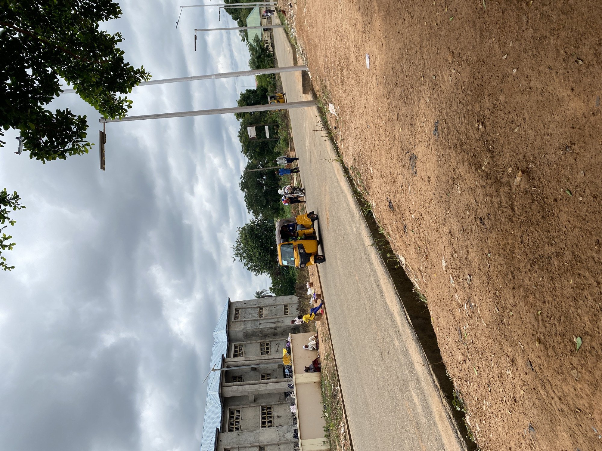 The image depicts a scene on a street in Lafia, Nigeria, under a cloudy sky. In the foreground, a reddish-brown dirt path lines the edge of an asphalt road. Along the road, several people are seated on the ground, some near a low wall. A yellow auto-rickshaw, also known as an 'Okada' or 'Keke', is visible driving on the road. In the background, a large building with a blue metal roof and multiple windows stands, appearing to be under construction or recently completed. Streetlights are positioned along the road, and trees and other vegetation are present. A few other individuals are walking on the road near the auto-rickshaw. The overall environment suggests an outdoor urban or semi-urban setting.
