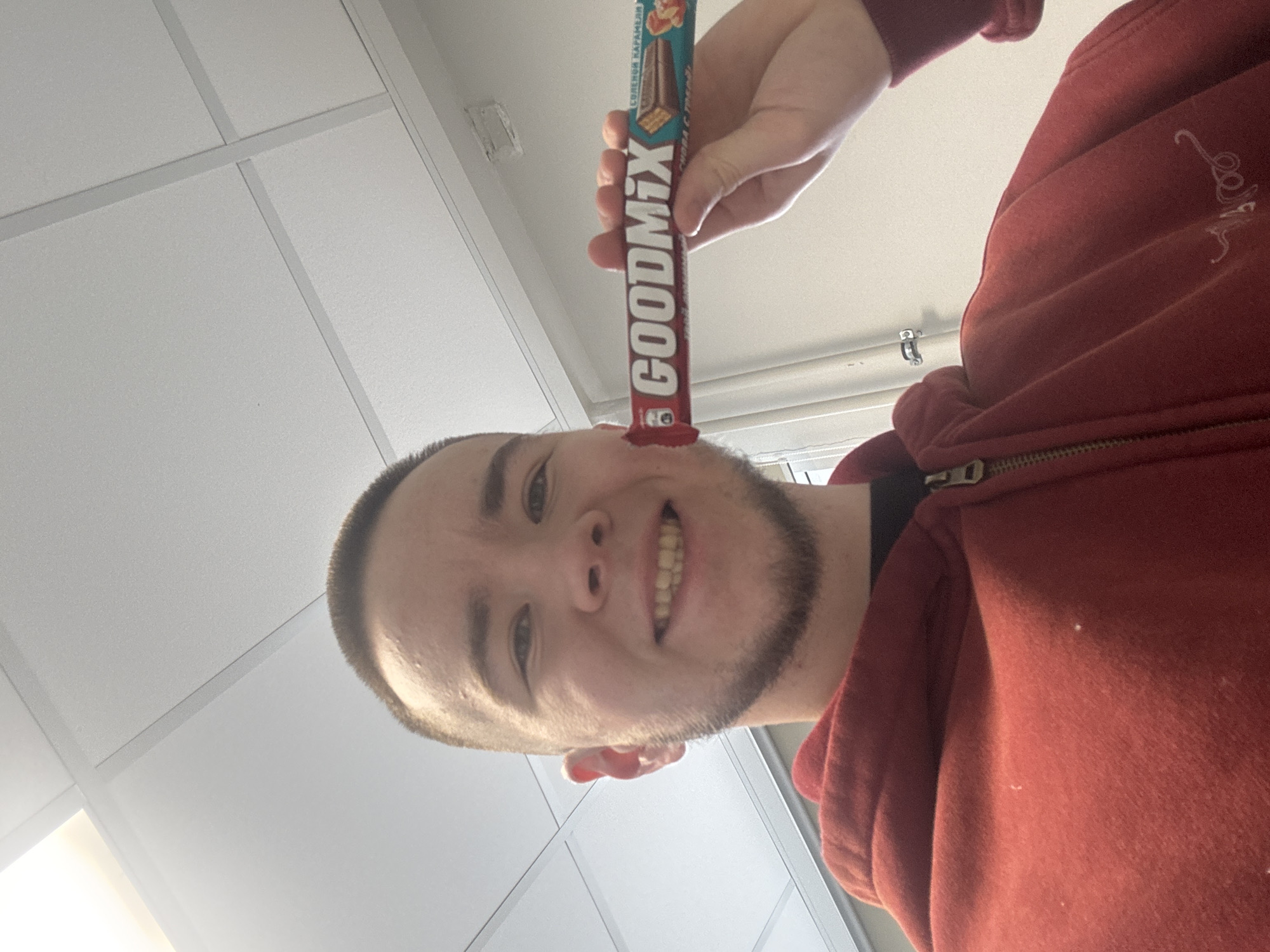 A young man is holding a "GOODMIX" candy bar vertically in front of his face, appearing to balance it on his nose. He is smiling, and the candy bar is oriented so the label is legible. He is wearing a red hooded sweatshirt. The background shows a typical suspended ceiling found in commercial or institutional buildings. The candy bar wrapper has Russian text indicating its origin. Although the image location is requested to be Kazan, Russia, the location cannot be determined from the given information.