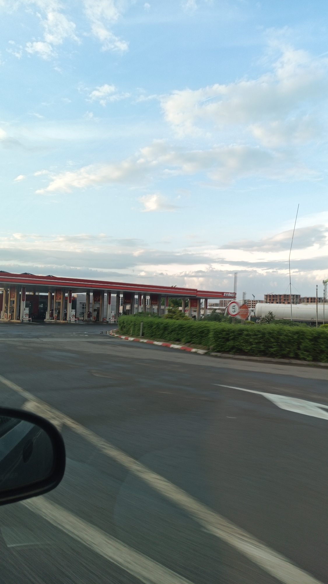 The image shows a gas station under a cloudy sky. The gas station has a long red roof. Green bushes are next to the road. A white arrow painted on the road points towards the right. A car's side mirror is visible in the bottom left corner. In the background, there is a white tank, possibly for gas storage, and unfinished construction buildings. A sign with the number 5 is also visible. Based on the "Лукойл" sign visible on the tanks, this is a Lukoil gas station. Given the architecture and features in the image, it is plausible that this gas station is located near Burgas, Bulgaria, potentially on a highway or road leading to/from the city.