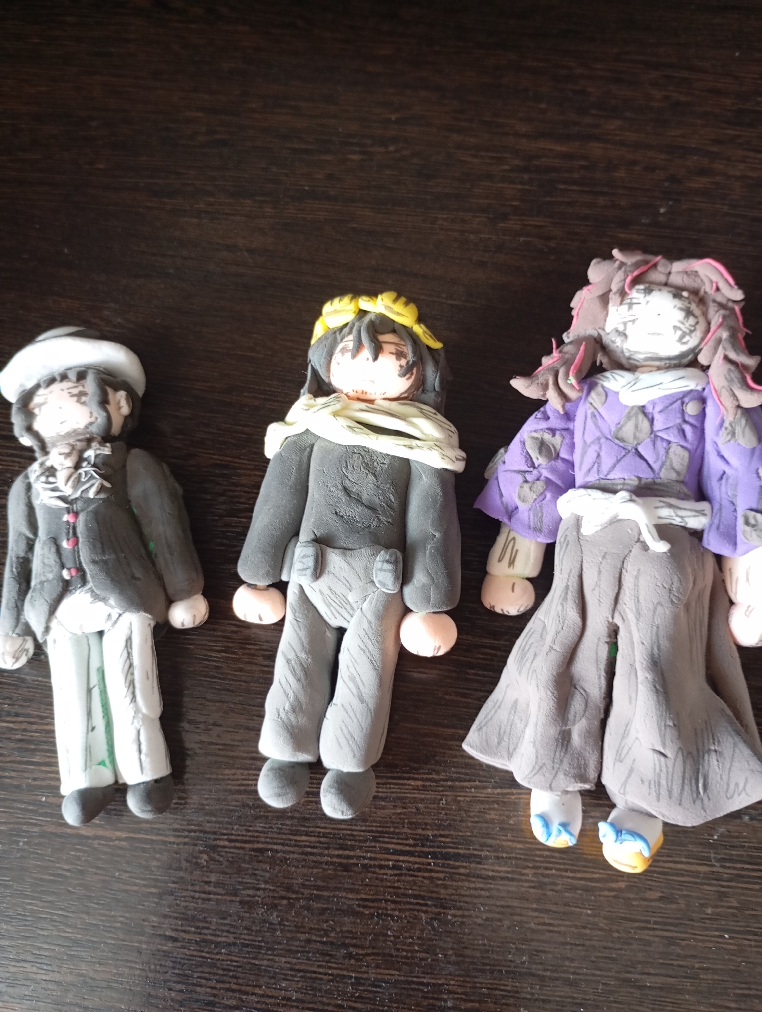 Three handmade clay figurines of anime-styled characters are displayed on a dark wooden surface. The figurine on the left wears a white hat, a dark coat with a white cravat, and striped pants. The middle figurine is dressed in a gray outfit with a yellow halo-like accessory around its head and a light-colored scarf. The figurine on the right sports a purple garment over a gray skirt, with pink accents in its hair and blue and yellow shoes. The figurines appear to be static, posed in a standing position. The background is a dark, textured wooden surface, likely a table. The image is taken from a slightly elevated angle, focusing on the figurines. There is no discernible location in the city of Льниха, Russia mentioned or depicted in the image.