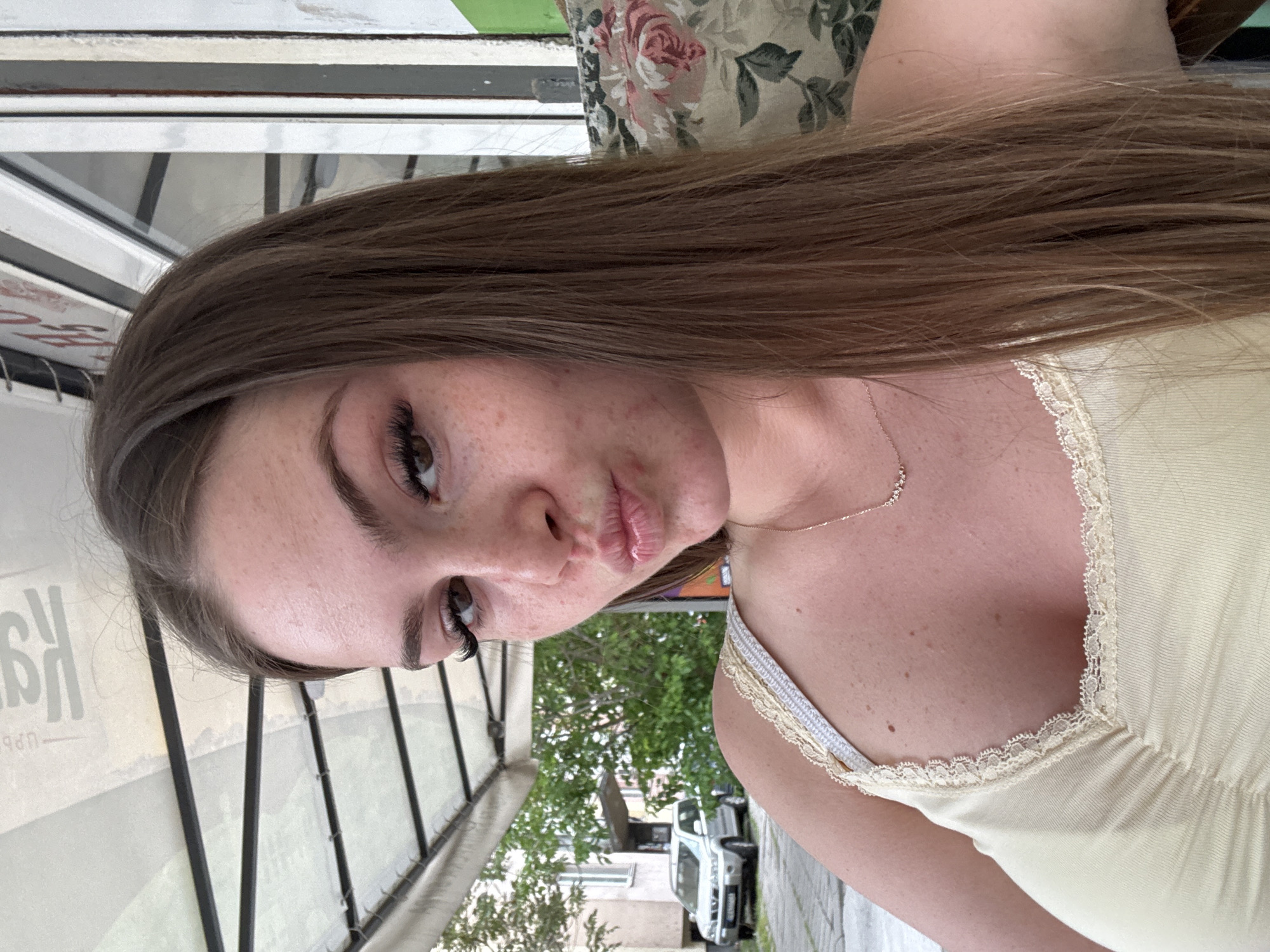 A woman is posing for a selfie outdoors. She has brown hair and is wearing a light yellow top with lace trim. A thin gold necklace is visible. She has light skin, freckles, and is making a kissing face. The background shows a sidewalk with trees, a white car parked on the street, and a building. The location appears to be in Plovdiv, Bulgaria, based on signage visible in the background with Cyrillic letters. Part of a covered outdoor seating area, possibly a restaurant or cafe, is seen to the left. The woman seems to be sitting or standing in this seating area. A floral patterned cushion or backdrop is seen behind her head.