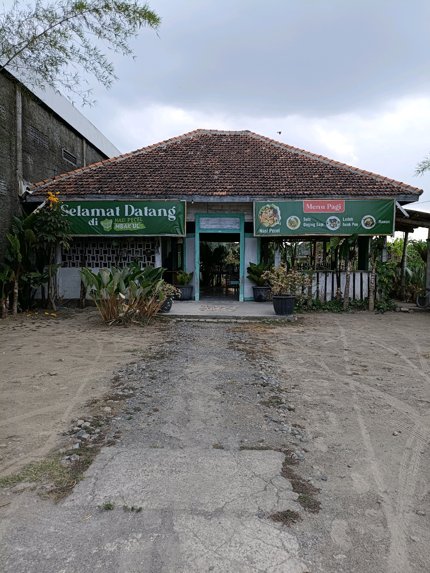 The image shows a small, traditional Indonesian restaurant located in Purwomartani, Indonesia. The restaurant has a rustic appearance with a tiled roof and an open-air seating area. A large green banner hangs above the entrance, displaying the words "Selamat Datang di Nasi Pecel Mbak Ul" which translates to "Welcome to Mbak Ul's Pecel Rice." Another banner to the right lists menu items such as "Soto Daging Sapi" (Beef Soup), "Lodeh Iwak Pee" (Fish Lodeh), and "Rawon" (a Javanese beef soup). The foreground is a dirt path leading to the restaurant, with some gravel and patches of grass. Several potted plants are placed around the entrance, adding a touch of greenery. The sky is overcast, suggesting a cloudy day.