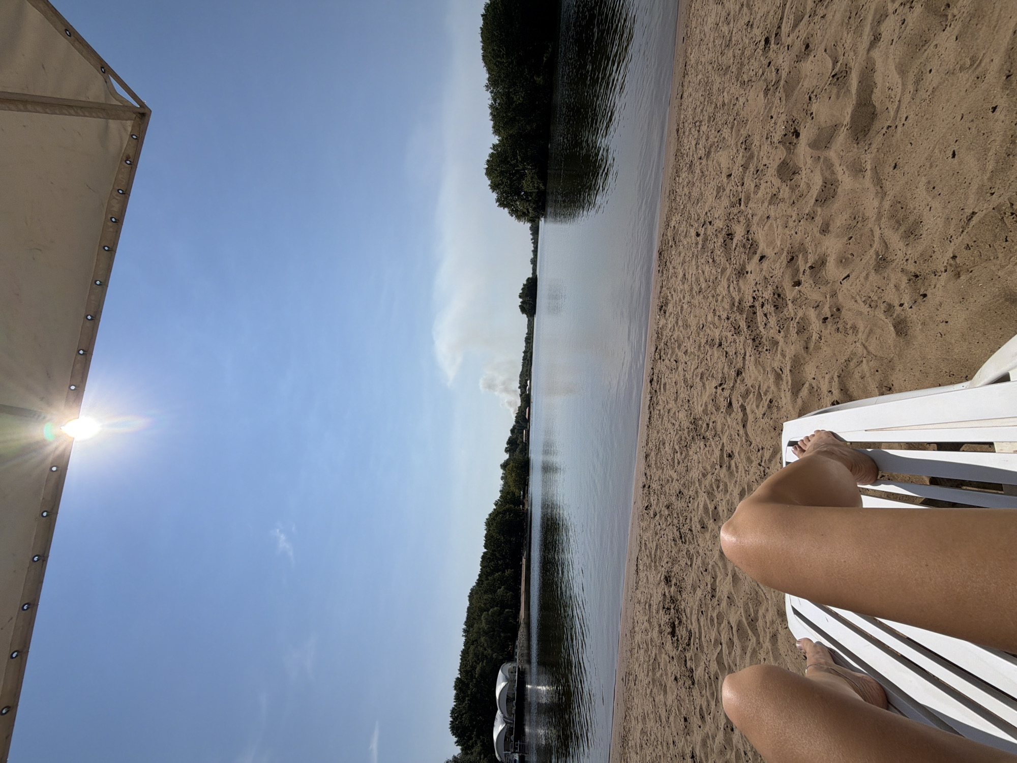 This is a first-person perspective photograph taken from a lounge chair on a sandy beach. The viewer's legs are visible in the foreground, resting on a white slatted lounge chair. The beach extends to the edge of a calm body of water. Across the water, a line of trees forms the distant shoreline, with a few buildings or structures visible among them. In the far distance, a plume of smoke rises into the bright blue sky. The sun is shining brightly, casting a glare in the upper portion of the image, partially obscured by a tan awning overhead. The location is identified as a beach in the city of Engels, Russia.