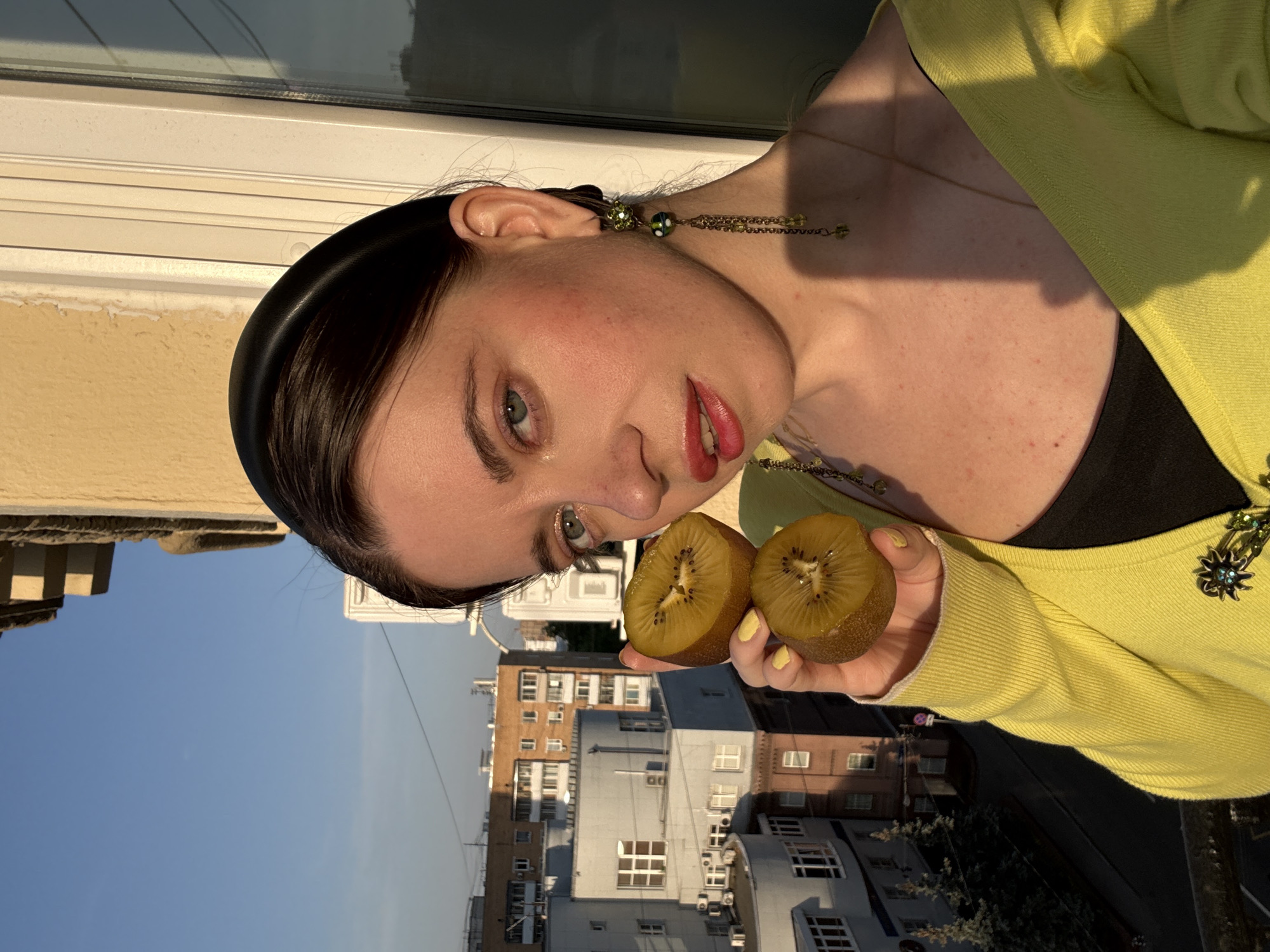 A woman with a black headband and green dangling earrings holds two halves of a golden kiwi fruit in her right hand. She wears a yellow cardigan over a black top and has red lipstick and painted yellow fingernails. The background features buildings typical of Moscow, Russia, under a clear blue sky, suggesting the photograph was taken outdoors, possibly on a balcony or rooftop. The location is determined by the distinctive architectural style of the buildings in the background.