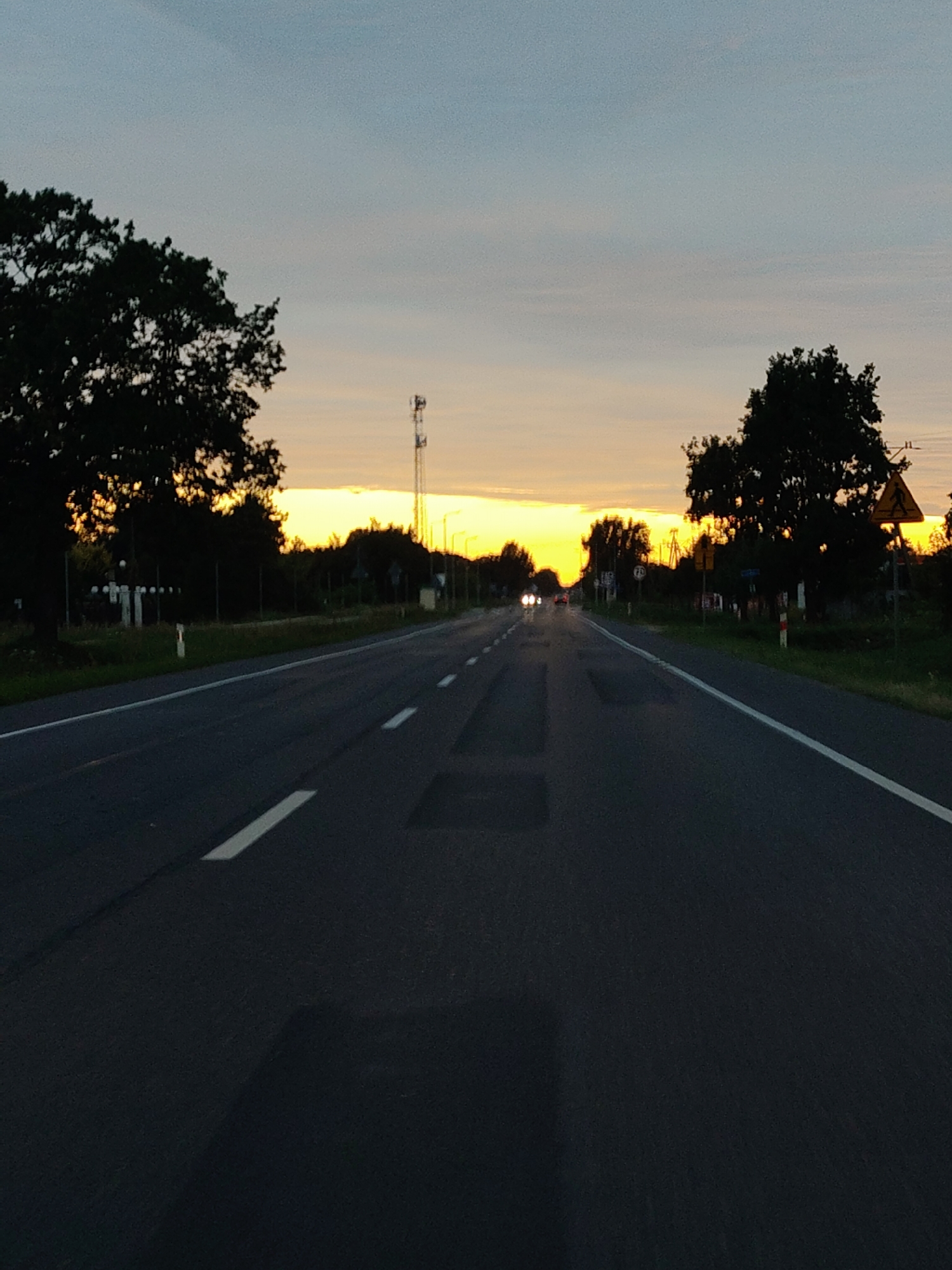 The image depicts a view down a rural road in Skierdy, Poland, at sunset. The road is asphalt and has white lane markings. On either side of the road, there are trees and vegetation. In the distance, the sky is a blend of orange, yellow, and blue, indicating the sun is setting. There is a cell tower visible on the left side of the road. There are also some road signs and streetlights visible. The image appears to have been taken from a moving vehicle.