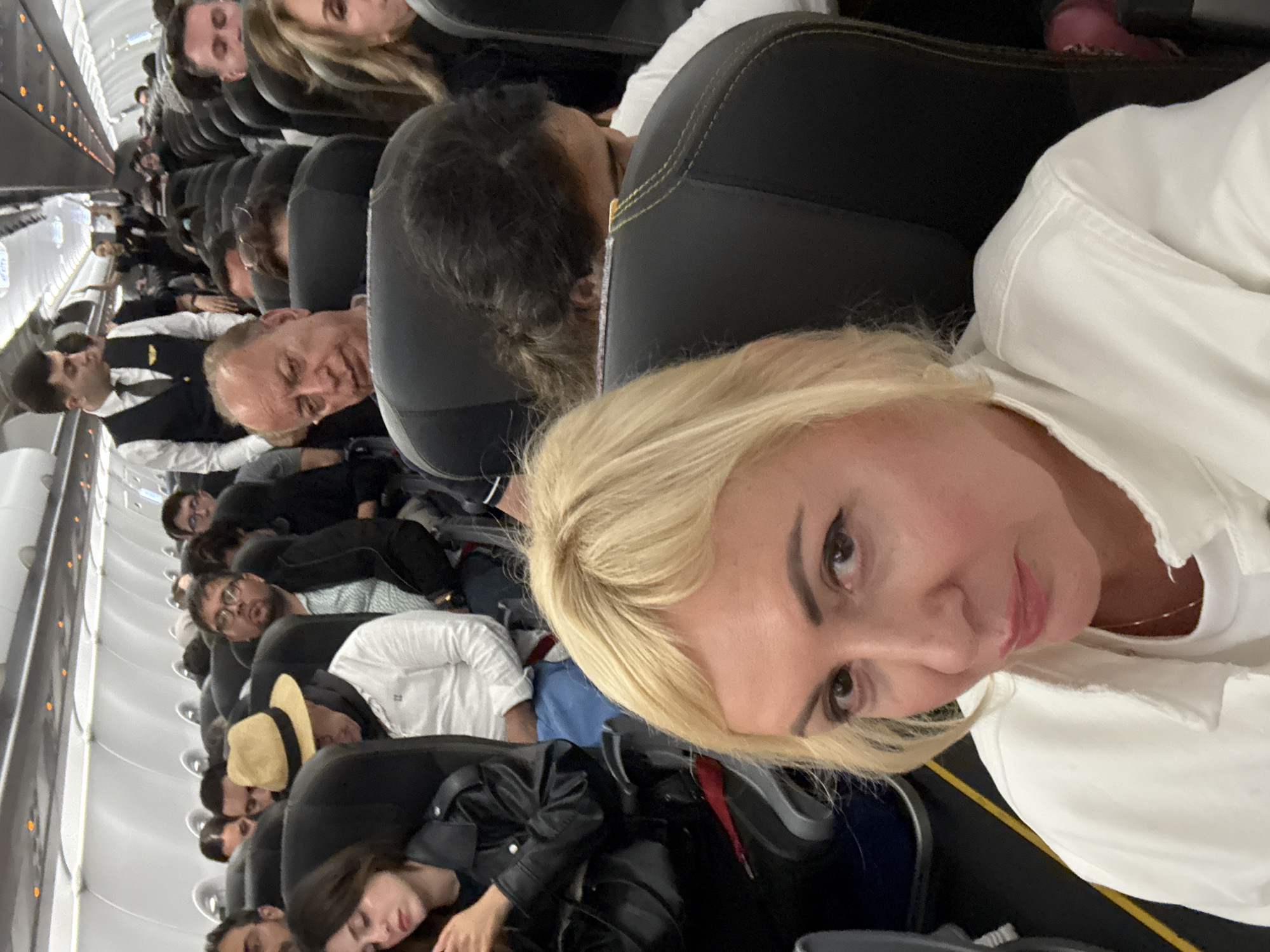 The image shows the interior of a crowded airplane cabin. Passengers are seated in rows of black seats, some sleeping or resting. One woman with blonde hair, wearing a white shirt, is taking a selfie. An airline employee in a black uniform and white shirt is visible further down the aisle. The airplane has white walls with an aisle running down the center.