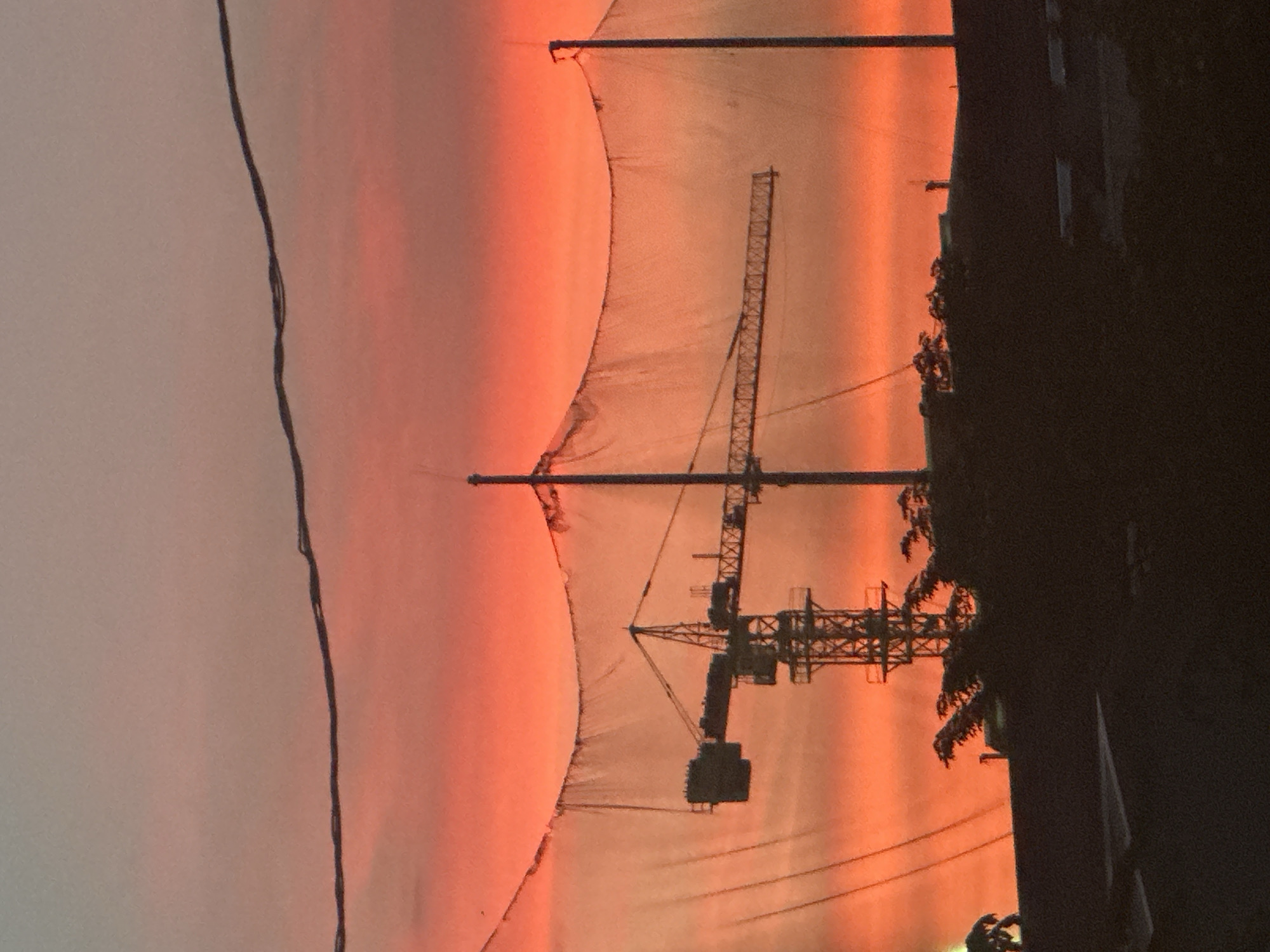 The image depicts a sunset in Da Nang, Vietnam, with a construction crane prominently featured against a vibrant orange and pink sky. The crane, a large piece of heavy machinery used in construction, is silhouetted against the colorful sunset. In front of the crane, a net is stretched between two poles, likely a safety net or a net used for sports or other activities. Below the crane and the net, a dark silhouette of buildings and trees can be seen, obscuring the ground and any potential activity occurring there. Overhead, electrical wires are visible, adding to the urban landscape element of the scene. The overall impression is one of urban development set against a dramatic natural backdrop.