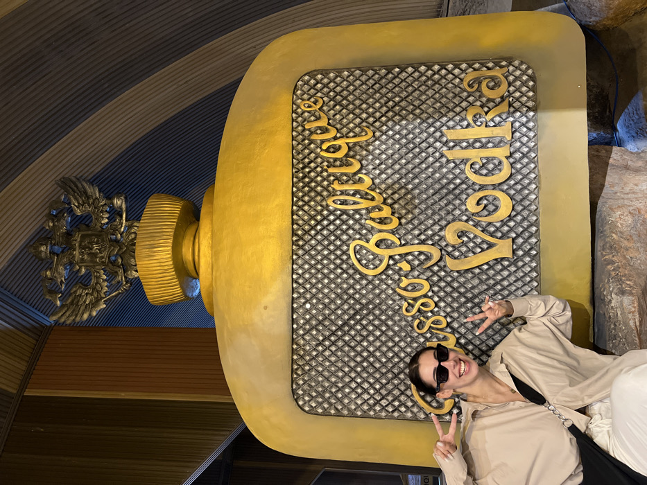 A woman with sunglasses poses with two peace signs next to a large, golden vodka bottle-shaped sign that reads "Beluga Vodka." The sign is textured with a diamond pattern on its surface. The bottle's cap features an ornate golden emblem. The background includes textured walls with blue and gray horizontal patterns, along with shades of brown. The woman is wearing a light-colored shirt and white pants.