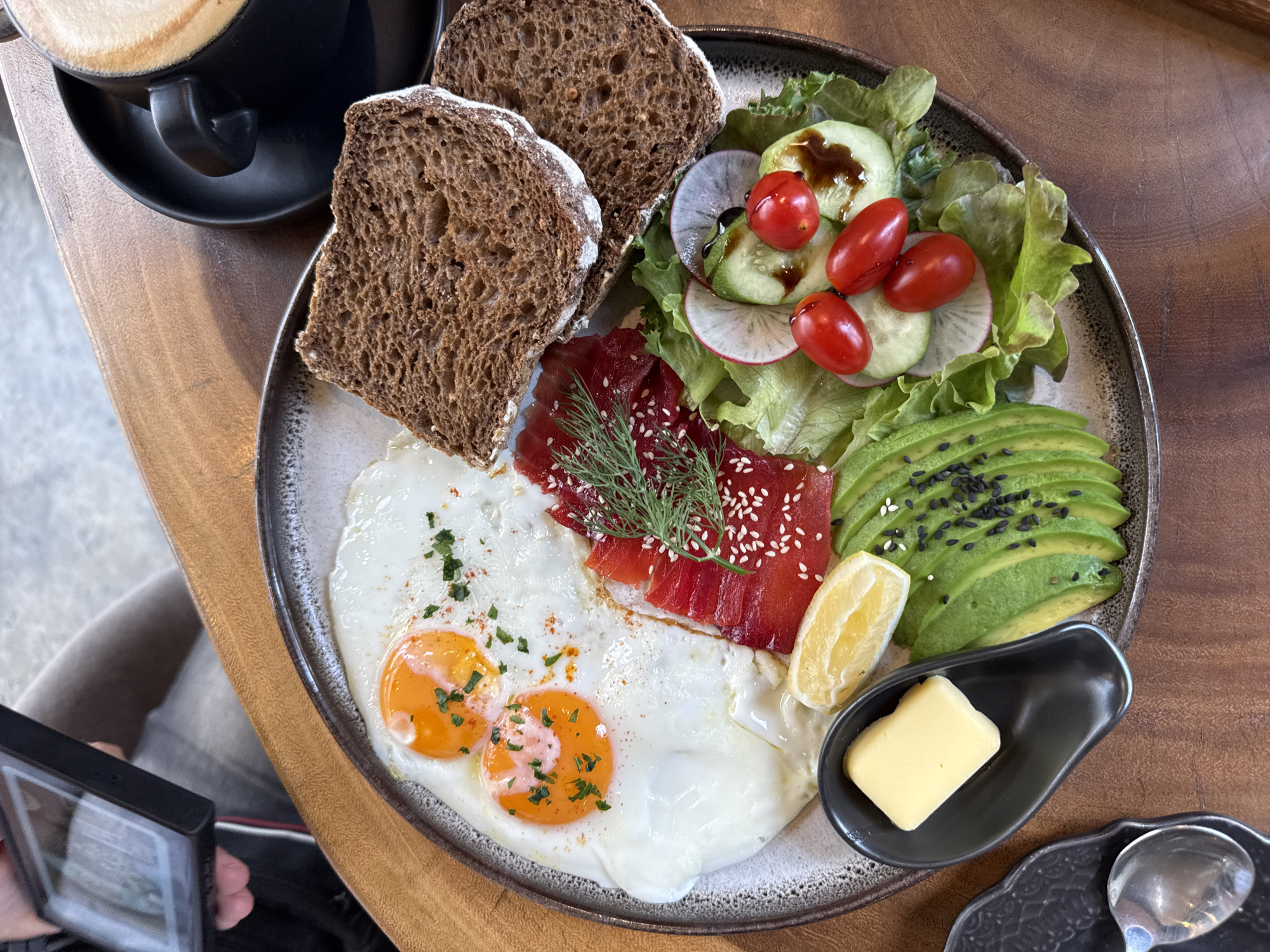 A close-up, overhead shot displays a breakfast spread on a wooden table in what appears to be Baan Bo Phut, Thailand. The primary subject is a circular plate filled with various breakfast items. Two sunny-side-up eggs with bright orange yolks are prominent, seasoned with chopped green herbs and a dusting of red spice. Beside the eggs, a portion of thinly sliced smoked salmon is artfully arranged, garnished with dill and white sesame seeds. To the right of the salmon, two slices of dark, textured bread sit next to a small salad composed of lettuce, cucumber slices, radish rounds, and cherry tomatoes drizzled with a dark sauce. A wedge of lemon is placed near the salmon, and a small, dark dish holds a pat of butter. To the lower left, a black dish with a spoon suggests another diner or preparation area. In the upper right corner, a black mug with a latte or cappuccino sits on a saucer, indicating a beverage is also part of the meal. A person's hand holding a smartphone is visible in the upper left, suggesting the food is being photographed. The lighting is natural and appears to be from an overhead source.