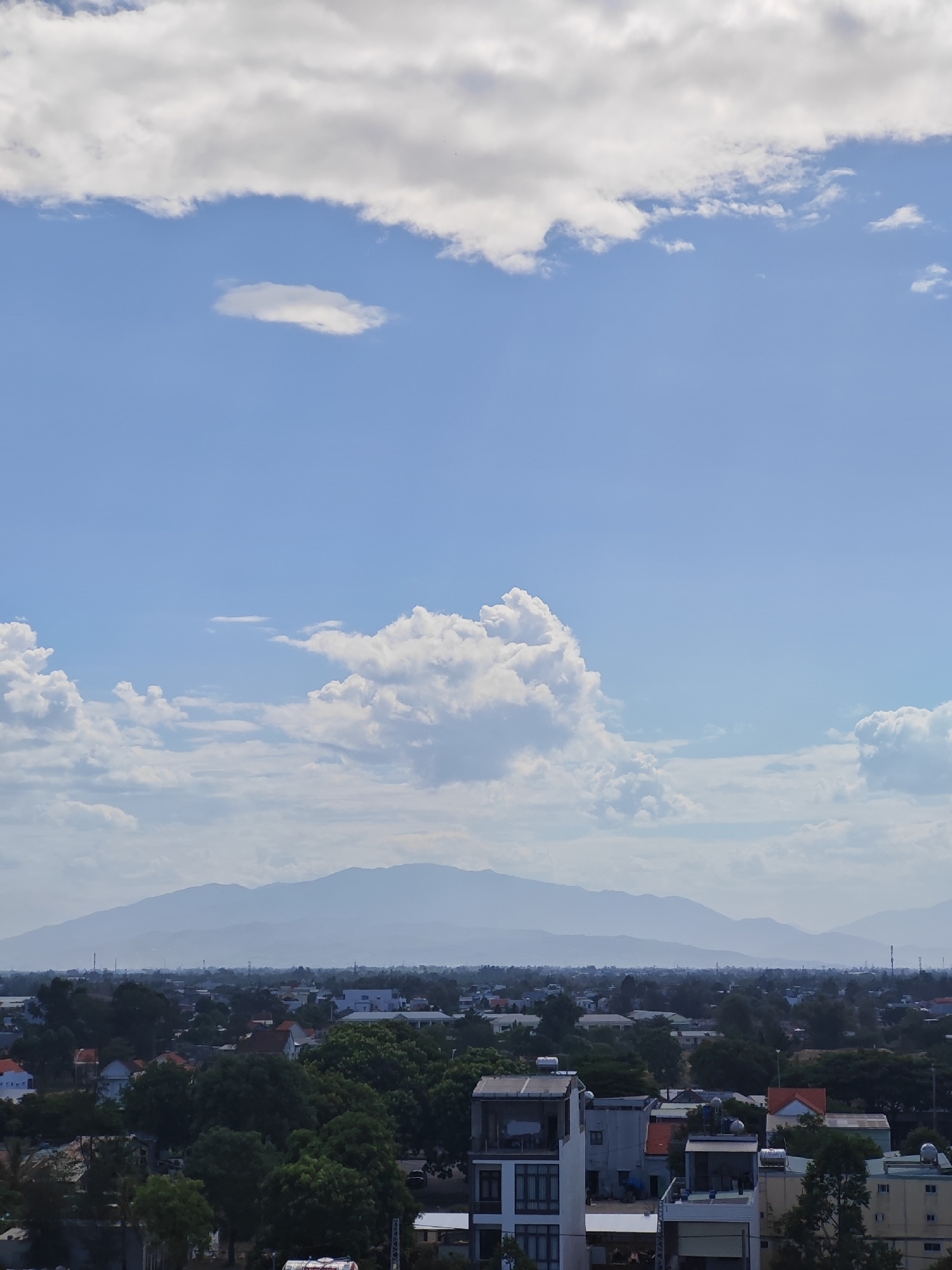 The image captures a panoramic view of a city with distant mountains under a partially cloudy blue sky. The foreground features a collection of buildings, ranging from multi-story structures to smaller residential homes, interspersed with dense green foliage. In the mid-ground, a vast urban sprawl unfolds, characterized by rooftops and scattered trees. The background is dominated by a series of hazy, layered mountains, suggesting a considerable distance. The sky above is a vibrant blue, with scattered white clouds, some appearing fluffy and voluminous, while others are thin and wispy. The lighting suggests it is daytime, likely mid-morning or afternoon, given the brightness and the shadows cast by the buildings.
This scene is consistent with a view from an elevated position within Đà Nẵng, Vietnam, looking inland towards the Trường Sơn mountain range. The architectural styles of the buildings, the prevalence of lush greenery, and the distinctive silhouette of the mountains are characteristic of the city and its surrounding landscape. The hazy appearance of the mountains is typical of atmospheric conditions in a tropical climate, where humidity and distance can soften distant features. There are no people or animals clearly visible in the frame, and no specific actions or interactions are depicted. The focus is on the urban landscape and its natural backdrop.
