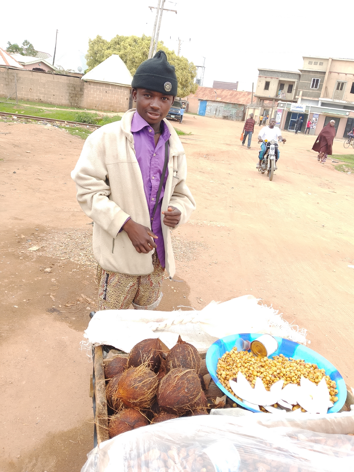 This media file shows a young man standing in front of a cart laden with coconuts and a bowl of fried snacks. He is wearing a purple shirt, a cream-colored jacket, and a black beanie with a circular emblem. His shorts have a patterned design. The cart is positioned on a dirt road, with a background that suggests a bustling street scene. In the distance, buildings with shops and residential units are visible. People are seen walking and riding a motorcycle, indicating daily life activity.
The time of day appears to be daytime, with overcast or hazy sky conditions. The presence of natural light suggests it's not nighttime. The setting seems to be an urban or semi-urban area, possibly a market or a busy street, given the shops and pedestrian traffic. The ground is made of dirt, and there are some signs of greenery and construction in the background.
Notable details include the way the young man is holding his jacket and looking towards the camera with a neutral expression. The cart is filled with whole coconuts, and a blue oval dish contains a pile of small, round, yellow fried snacks, possibly chickpeas or a similar item, accompanied by some white cutouts that resemble flower petals, likely made from coconut. A small can of oil or a similar product is also present in the dish. Some plastic sheeting is used to cover parts of the cart's contents.
There is some text visible on the shops in the background, including what appears to be "Merdos" and "ROS" on one of the storefronts. The man's shorts also have some lettering on them, though it's difficult to read clearly.