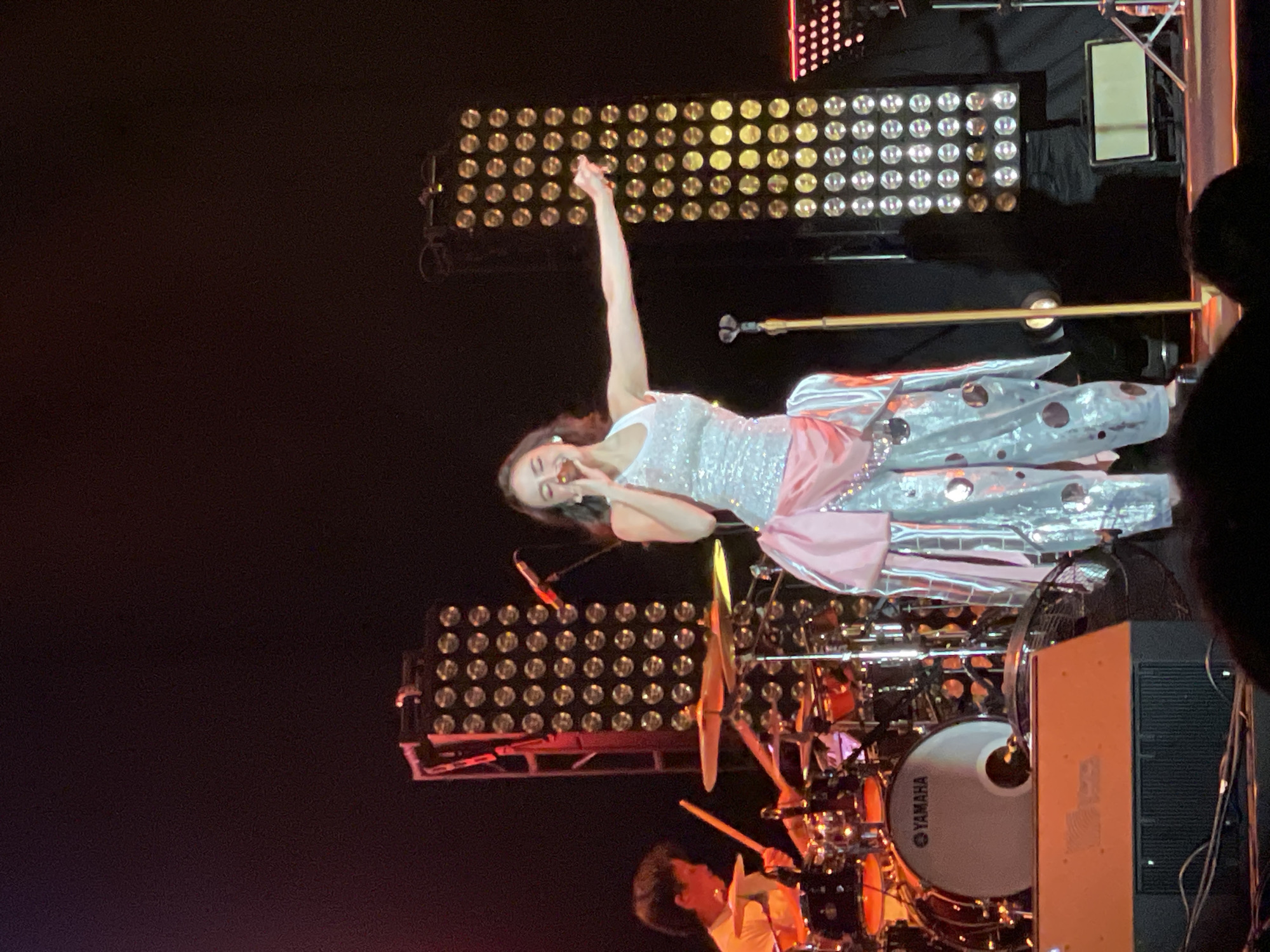 The image depicts a female singer performing on stage. She is wearing a sparkly top and a pink and silver skirt, with one arm raised in the air. She is holding a microphone and appears to be singing. Behind her is a drummer playing a Yamaha drum set. Stage lights are visible in the background. The location is likely Salembaran, Indonesia, based on the model's usual performance location.