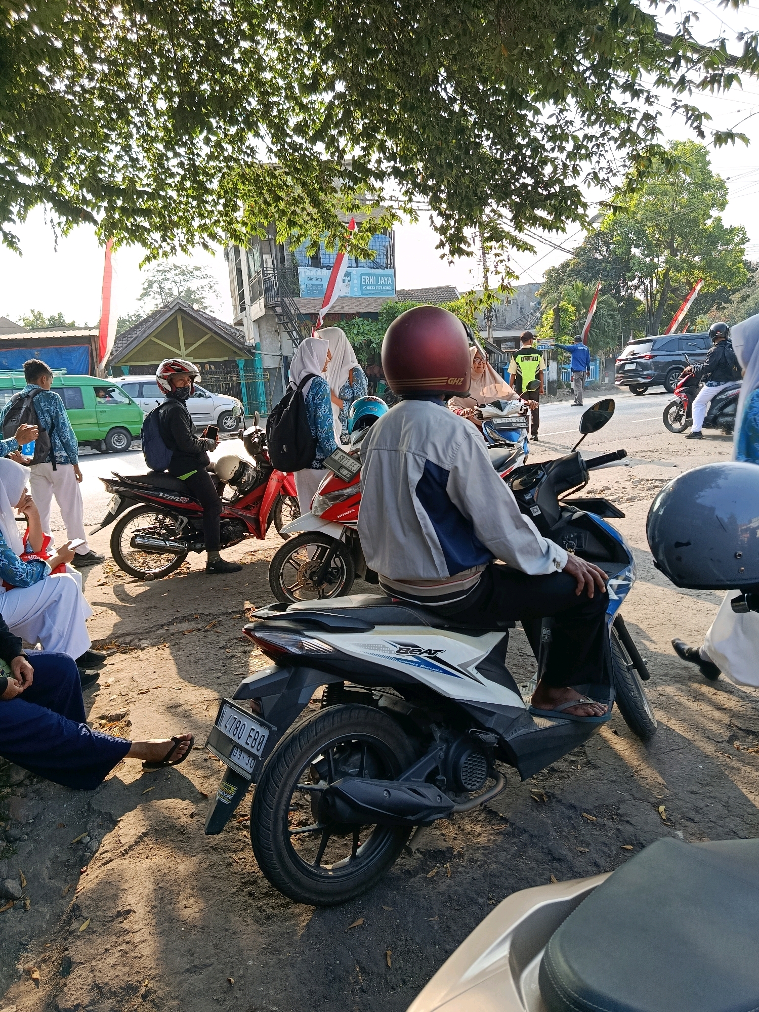 The image captures a street scene in Bululawang, Indonesia, under the dappled sunlight filtering through tree branches. In the foreground, a person wearing a red helmet and a grey and blue jacket is seated on a white and blue motorbike. The motorbike has a license plate that reads "N 4780 EBO" with a sticker below showing "09.30". To the left of this motorbike, another red motorbike is parked, with a person wearing a red helmet and a dark jacket seated on it, holding a mobile phone.
Several individuals in school uniforms, consisting of blue patterned shirts and white pants, are present. Some are standing near the motorbikes, while others are seated on the ground. A green van is parked in the background, and a dark SUV is visible on the road. Two Indonesian flags, red and white, are displayed on poles, one attached to a building and another on a separate pole. The environment appears to be a road with a dirt and gravel shoulder, bordered by buildings and lush green trees. The overall atmosphere suggests a moment of pause or waiting, possibly before or after school.